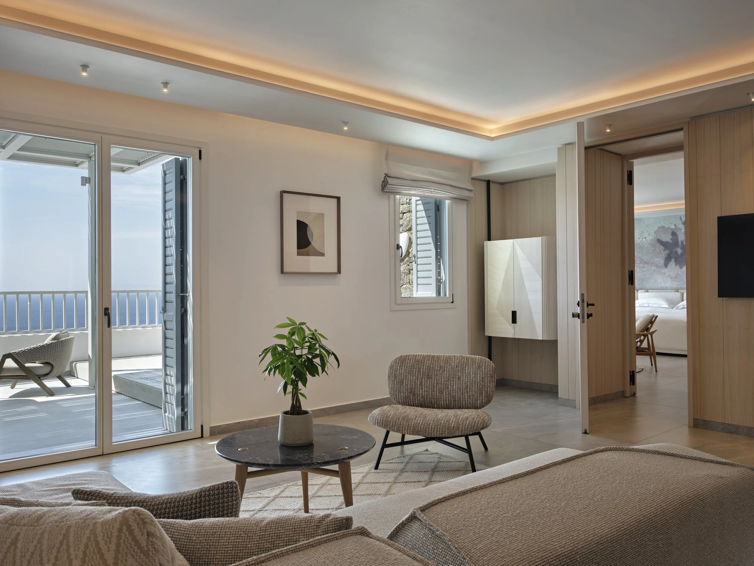 Living room with sliding glass door leading to balcony, a beige sofa, a small round coffee table with a potted plant, a patterned armchair, and a flat-screen TV on the wall. The interior features minimalist decor, light wood accents, and a view of th