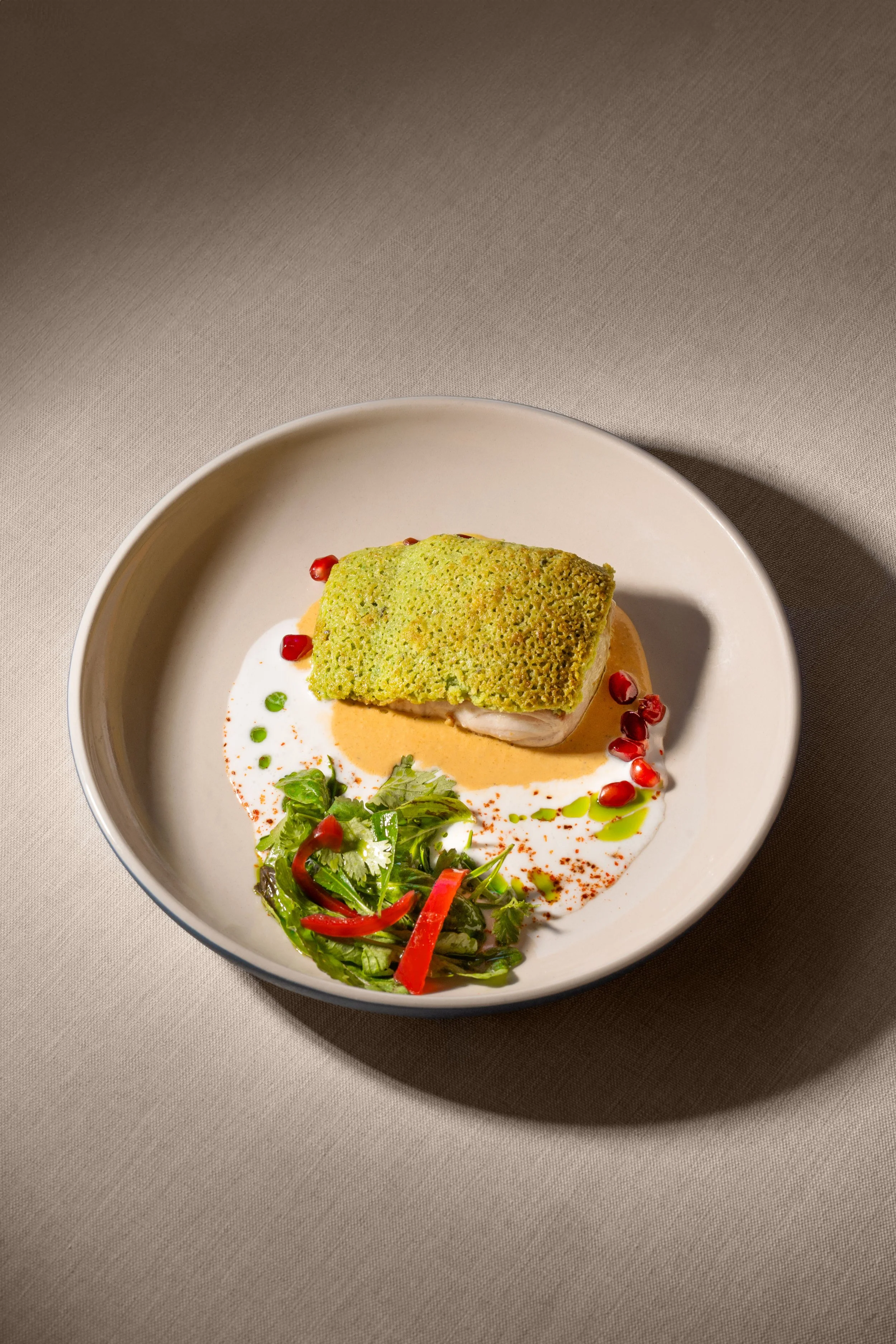 A plated gourmet dish featuring a piece of fish topped with a crispy green crust, garnished with pomegranate seeds, a small fresh salad, and drizzled sauces on a beige plate.