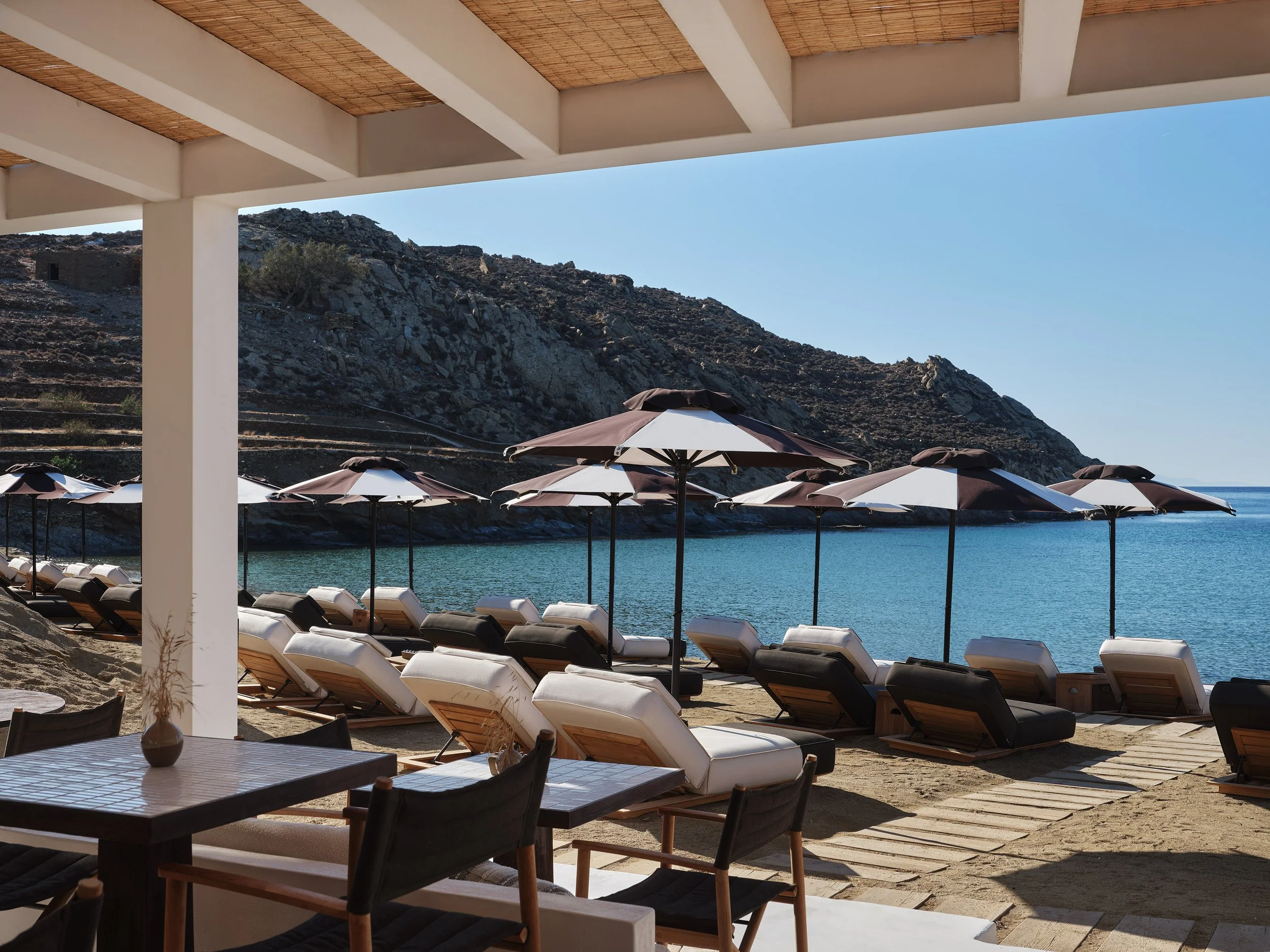 Beachside lounge chairs with umbrellas on sandy beach, view of calm water and rocky hillside in the background, under a shaded patio area.