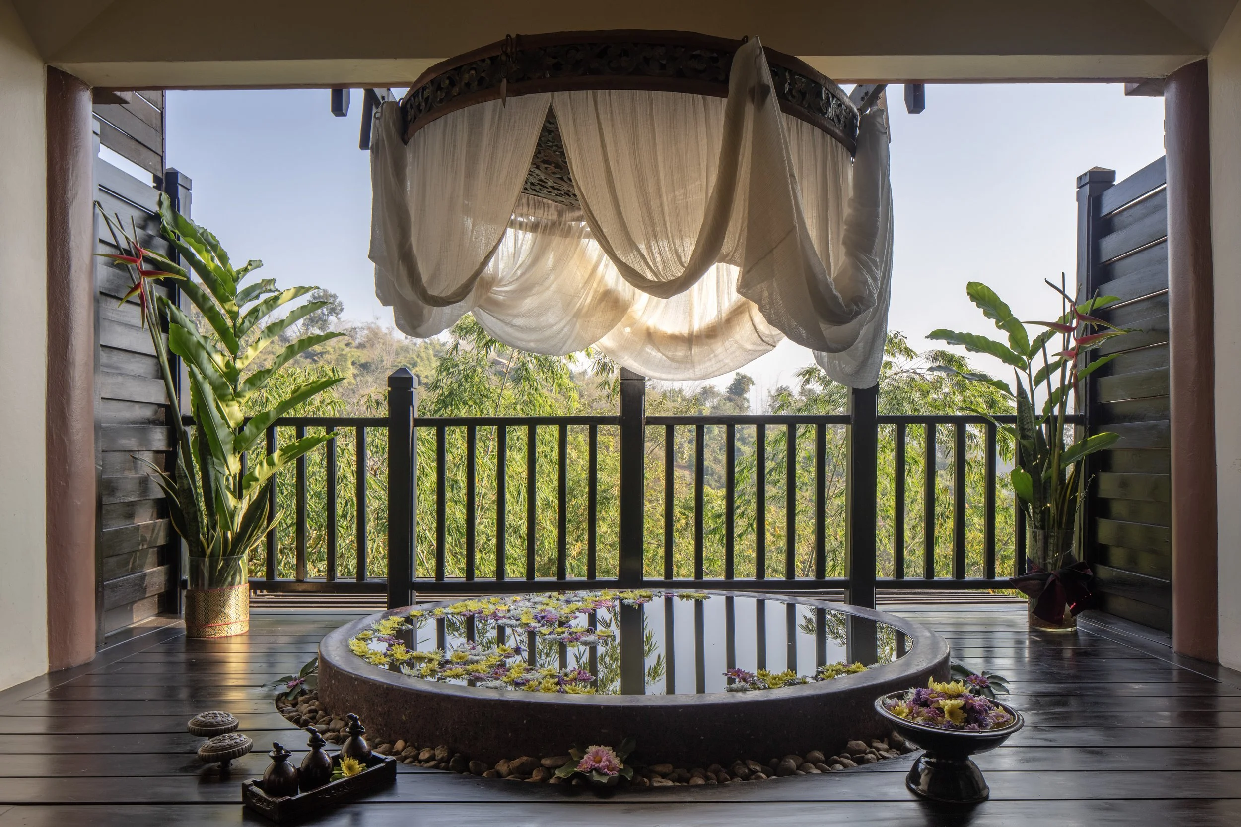 Balcony with a small hot tub filled with floating flowers, two potted plants on either side, a hanging white curtain, and a scenic view of trees and hills outside.