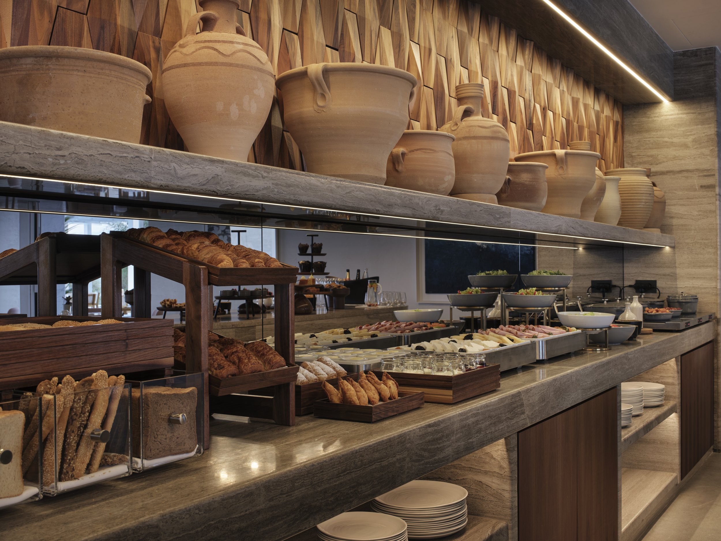 Buffet table with baked goods, salads, and cold cuts, with large pottery vases on wood-panelled wall in background.