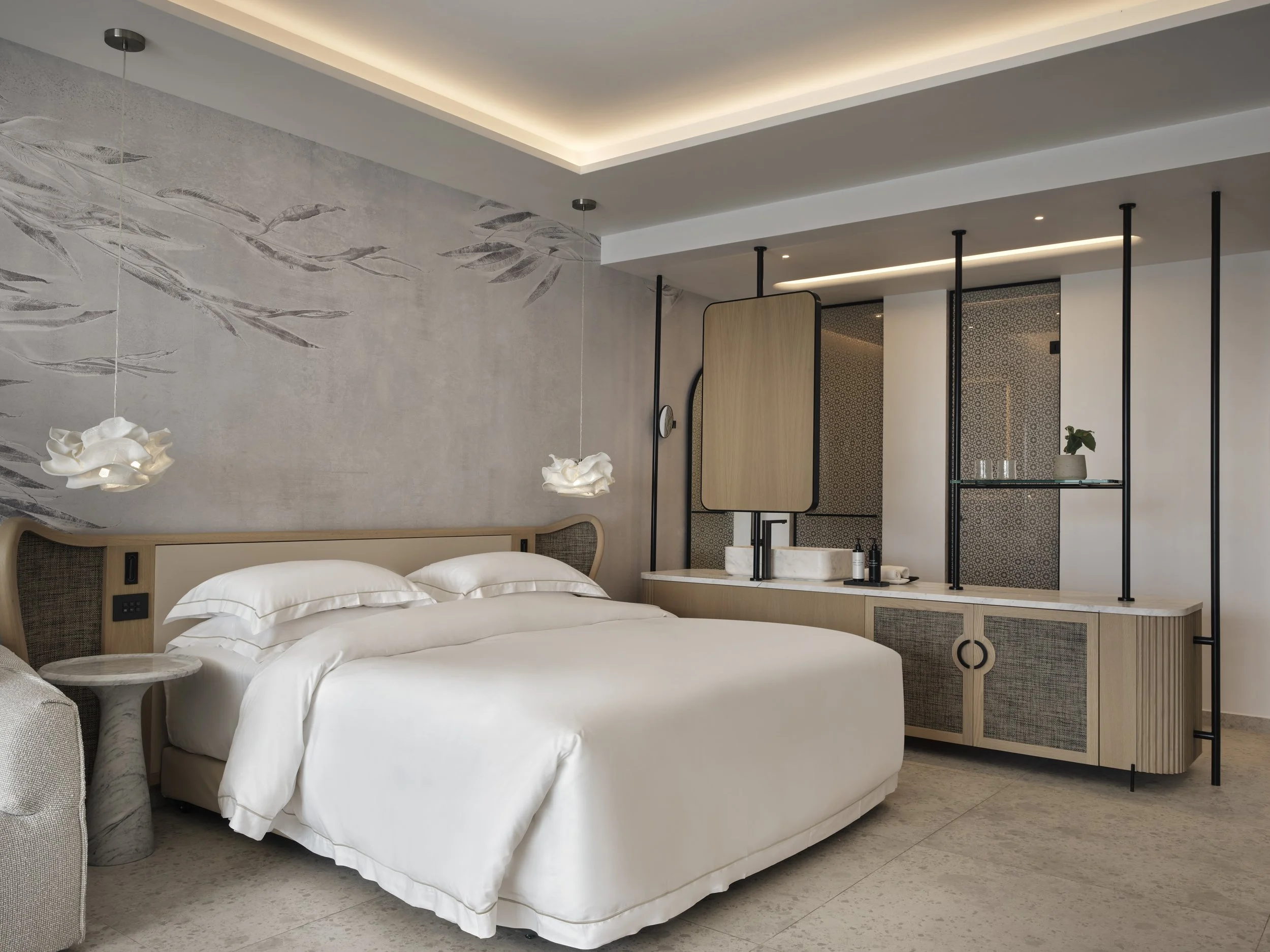 Modern hotel room with a white bed, wall art, pendant lights, and a wooden divider with decorative shelving, a sink, and a small potted plant.