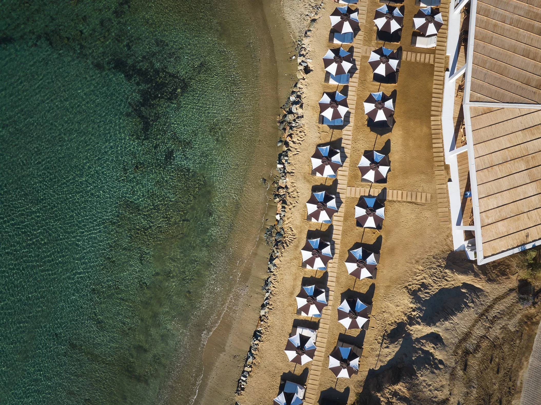 An aerial view of a beach with two rows of white umbrellas, sandy shore, clear green water, and a building with a wooden roof on the right side.