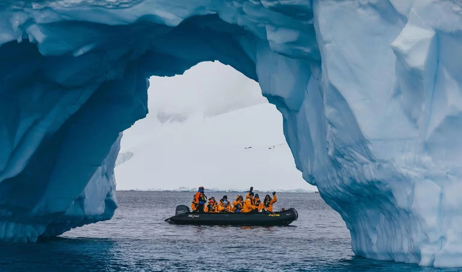 Group of people in orange jackets on a black inflatable boat exploring icy waters, framed by a large ice cave arch with snowy landscape in the background.