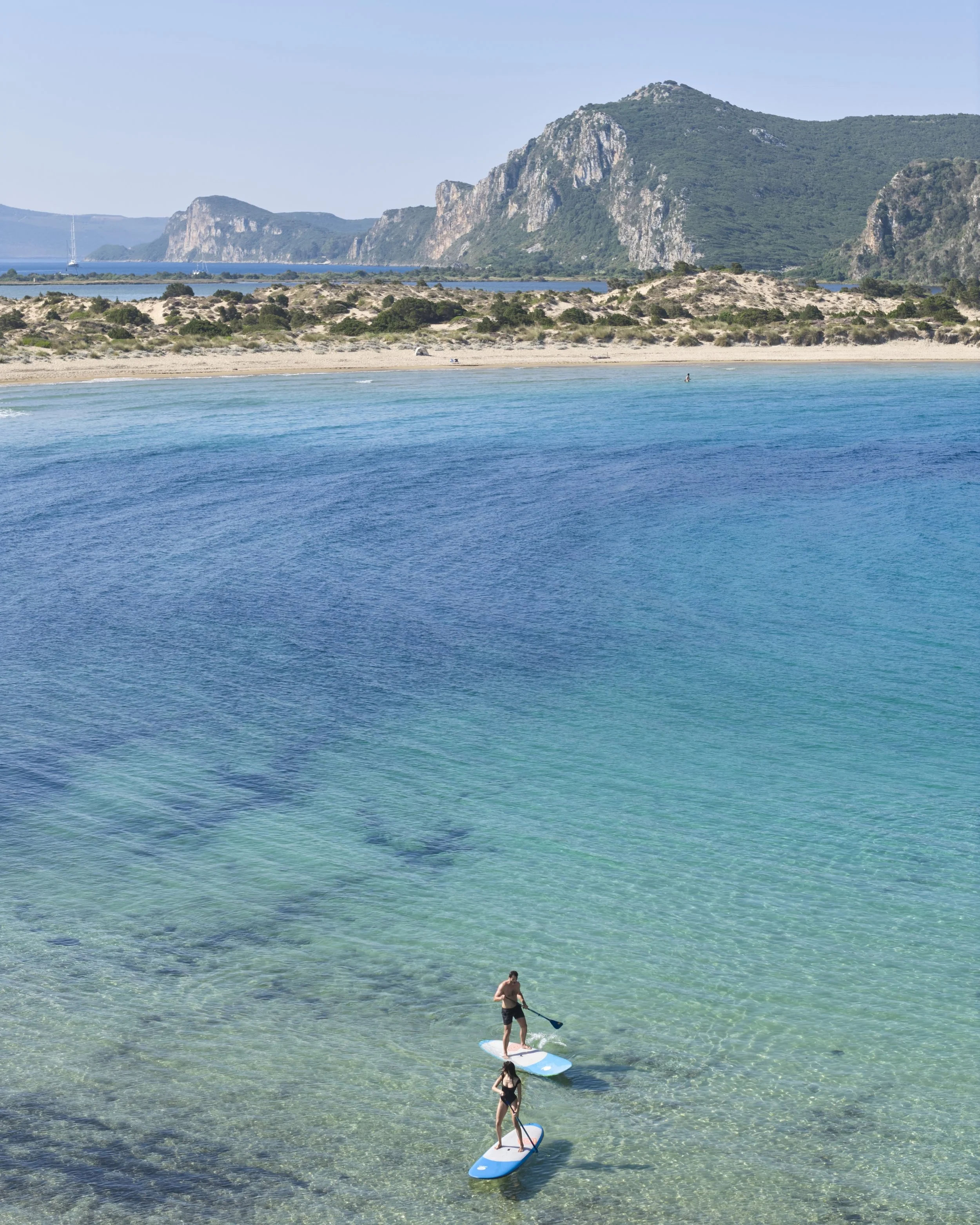 Three people paddleboarding in clear, turquoise water near a sandy beach with rocky hills and cliffs in the background.