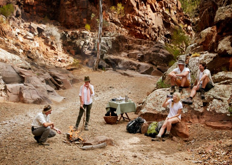 Group of people camping and relaxing in a rocky outdoor canyon area with a small campfire, table, and backpack.