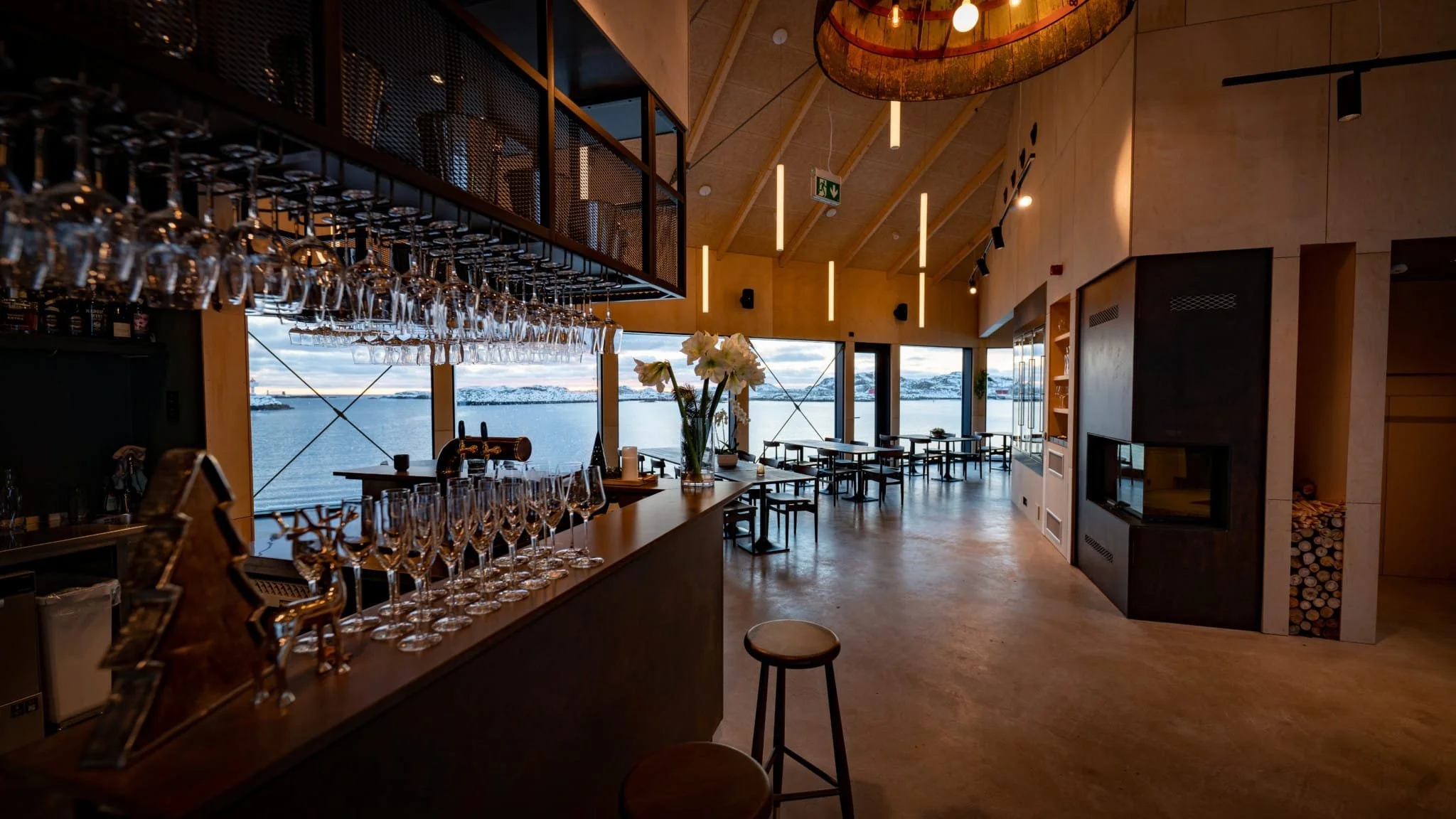 Interior of a modern restaurant or bar with large windows overlooking a body of water and snow-capped mountains in the distance. The space features a high wooden ceiling, hanging lights, a bar area with glassware, and a central vase of white flowers.