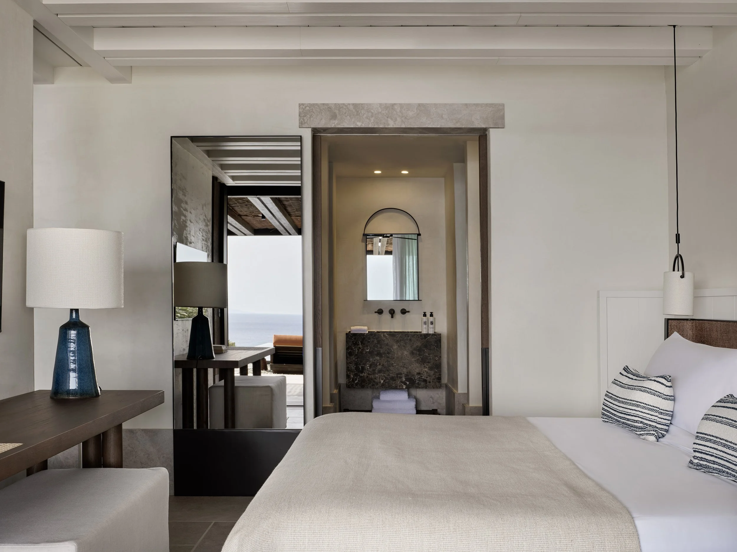 A hotel room with a bed, pillows, bedside lamps, a mirror, and a view of an outdoor area and ocean through open doors.