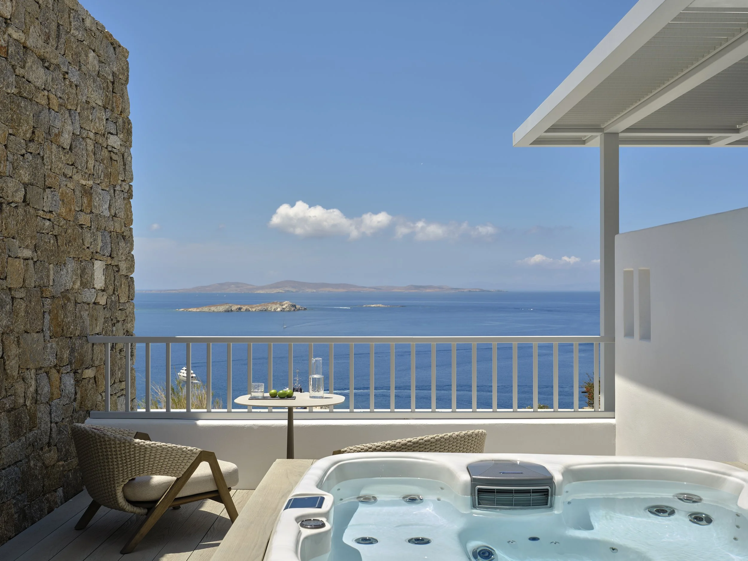 Balcony with jacuzzi overlooking the ocean, with a small table and chairs, stone wall, and white railing, under a blue sky with clouds.