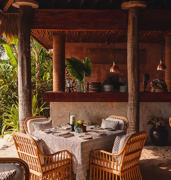 Outdoor dining area with a table set for four, surrounded by wicker chairs with cushions, under a rustic wooden roof with hanging pendant lights, and lush tropical plants in the background.