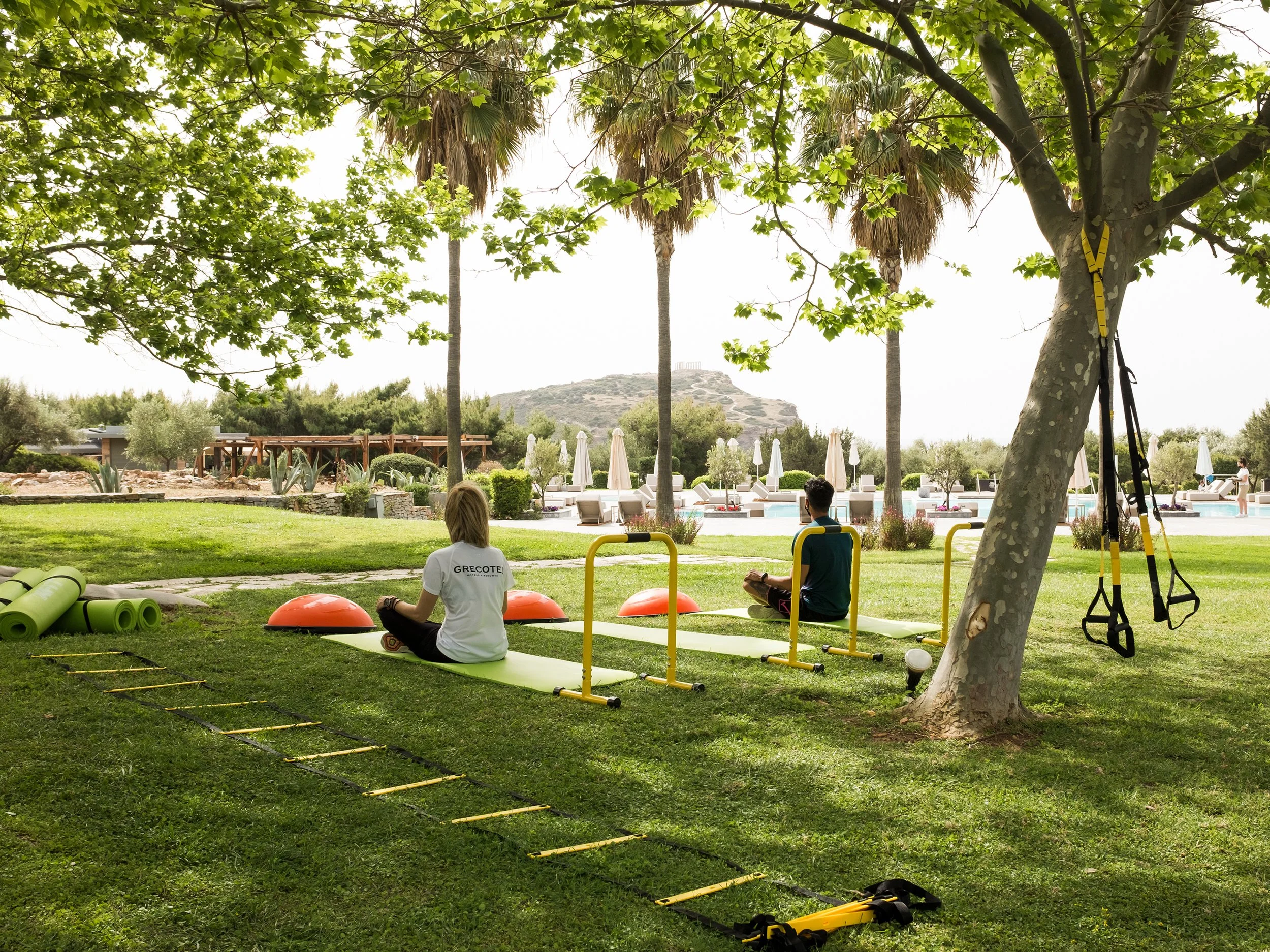 Two people sit on yoga mats in a shaded outdoor park, practicing meditation or yoga, with workout equipment and a swimming pool in the background.