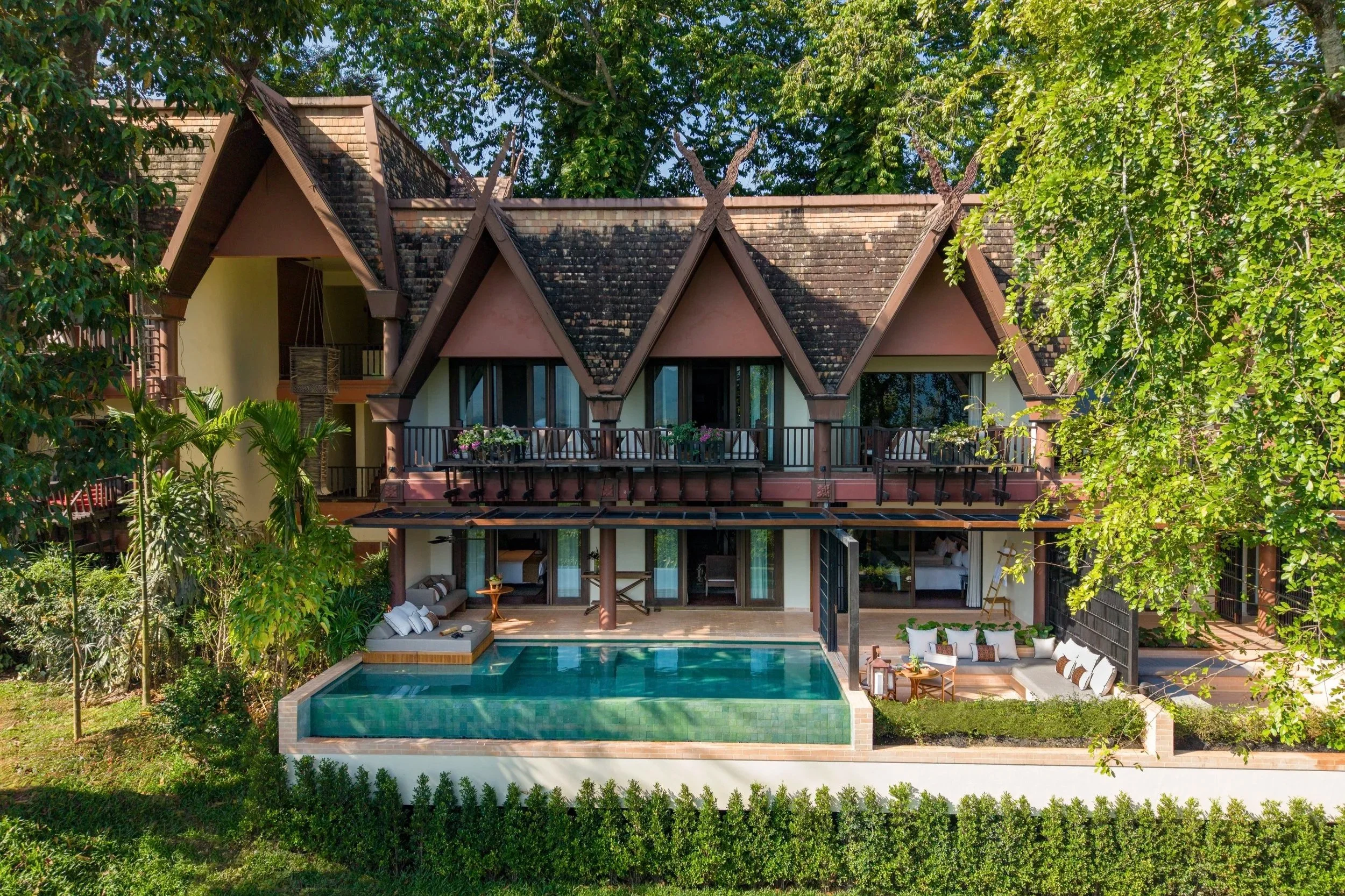 A luxurious two-story house with a tiled roof, balconies, and large windows surrounded by lush green trees. The backyard features a swimming pool, outdoor lounge area, and patio furniture.