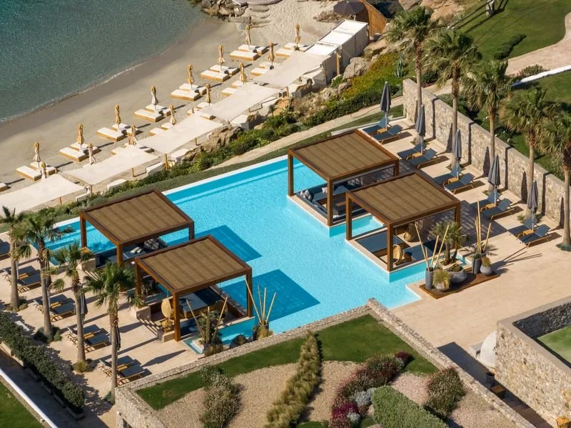 Luxury pool area with three shaded cabanas, surrounded by lounge chairs, umbrellas, and palm trees, overlooking a beach with sunbeds and umbrellas.