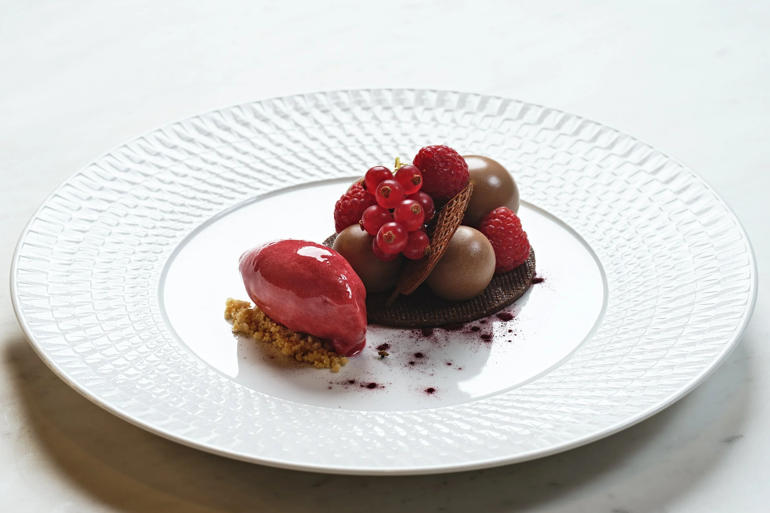 Elegant plated dessert with chocolate spheres, red berries, a red sorbet, and decorative chocolate wafer on a white textured plate.