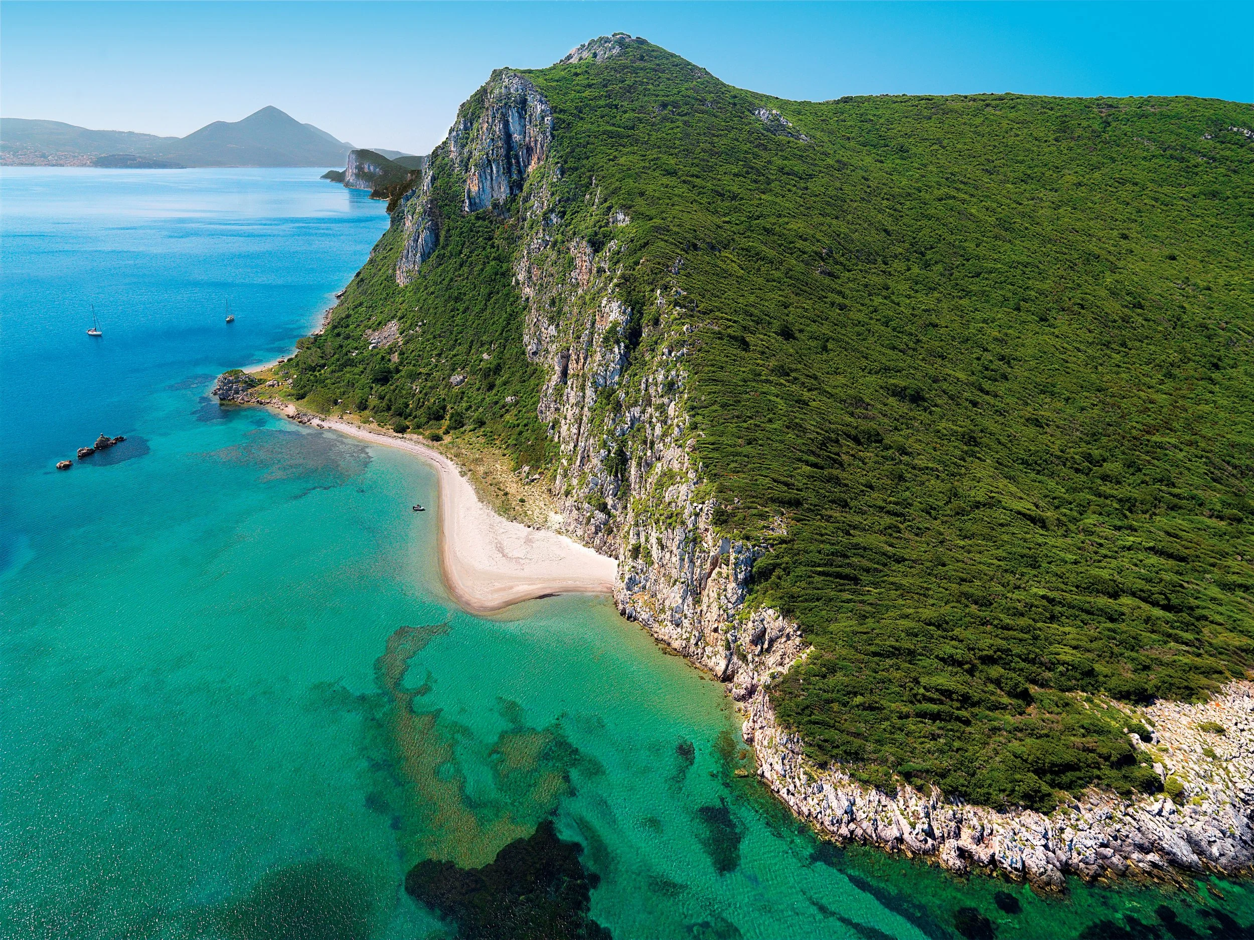 Aerial view of a green mountain overlooking a curved sandy beach and turquoise sea with boats, under a clear blue sky.