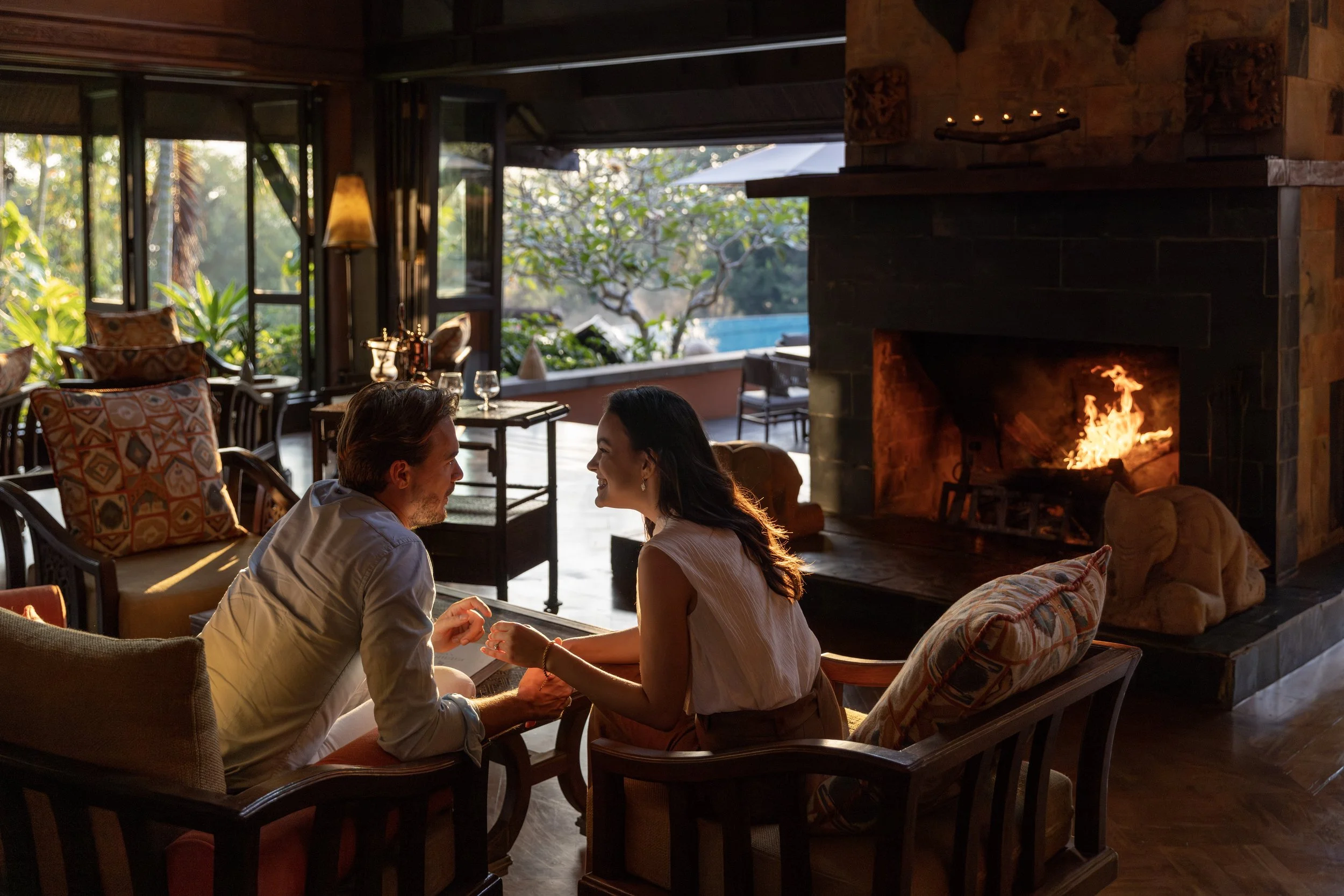 A couple sitting by a fireplace in a warmly lit living room, holding hands, with an outdoor view of greenery and a pool in the background.
