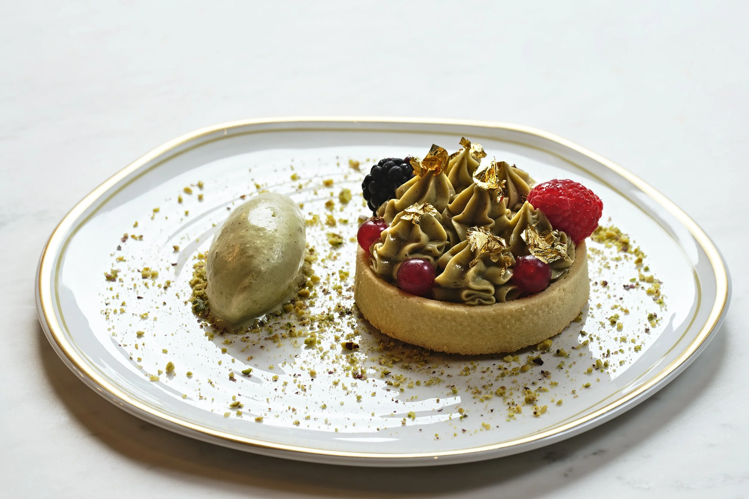 An elegant dessert on a white plate with gold trim, featuring a circular tart topped with piped greenish-brown cream, fresh berries, edible gold leaf, and a side quenelle of green ice cream, garnished with crushed pistachios.