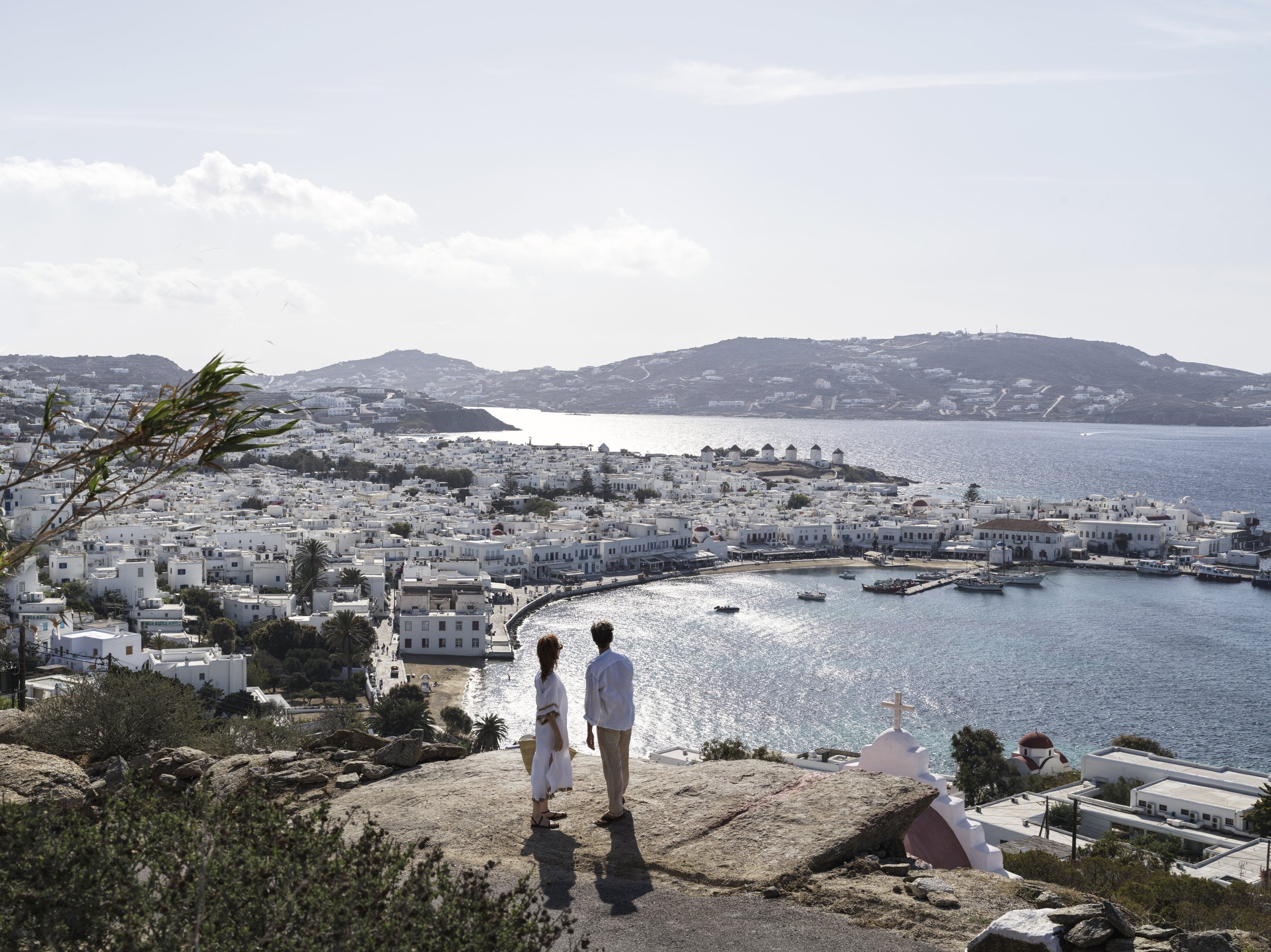 A man and woman holding hands and talking, standing on a rocky hill overlooking a coastal town with white buildings, a harbor, and mountains in the distance on a bright day.
