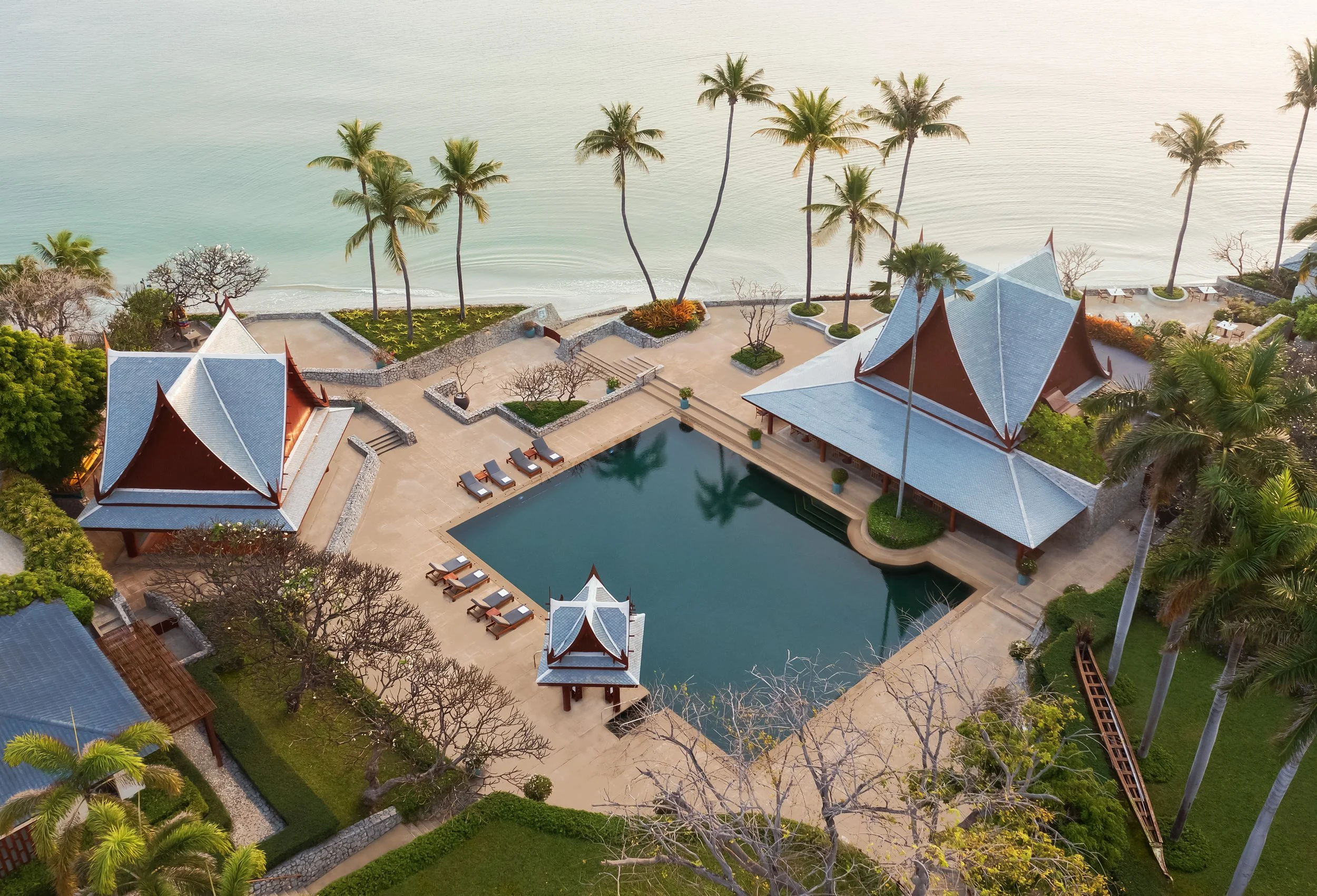 Aerial view of a luxurious beachfront resort with traditional Thai-style pavilions, a swimming pool, palm trees, and lounge chairs overlooking the ocean.