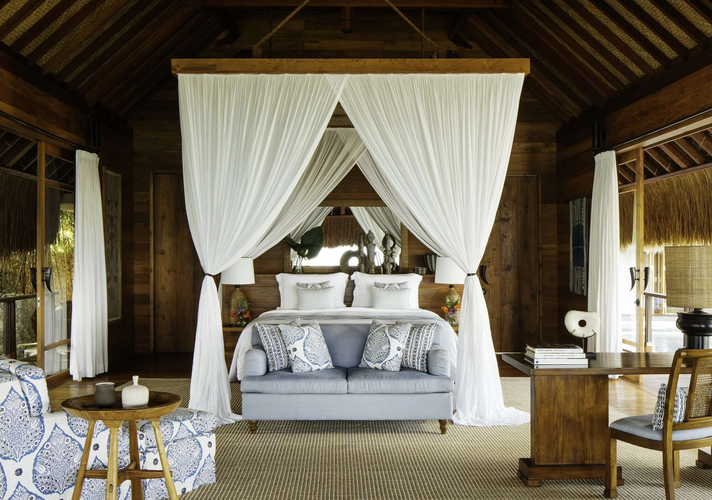 A bedroom with a four-poster bed, white curtains, a blue sofa with patterned pillows, a round wooden table with vases, and wooden walls and ceiling.