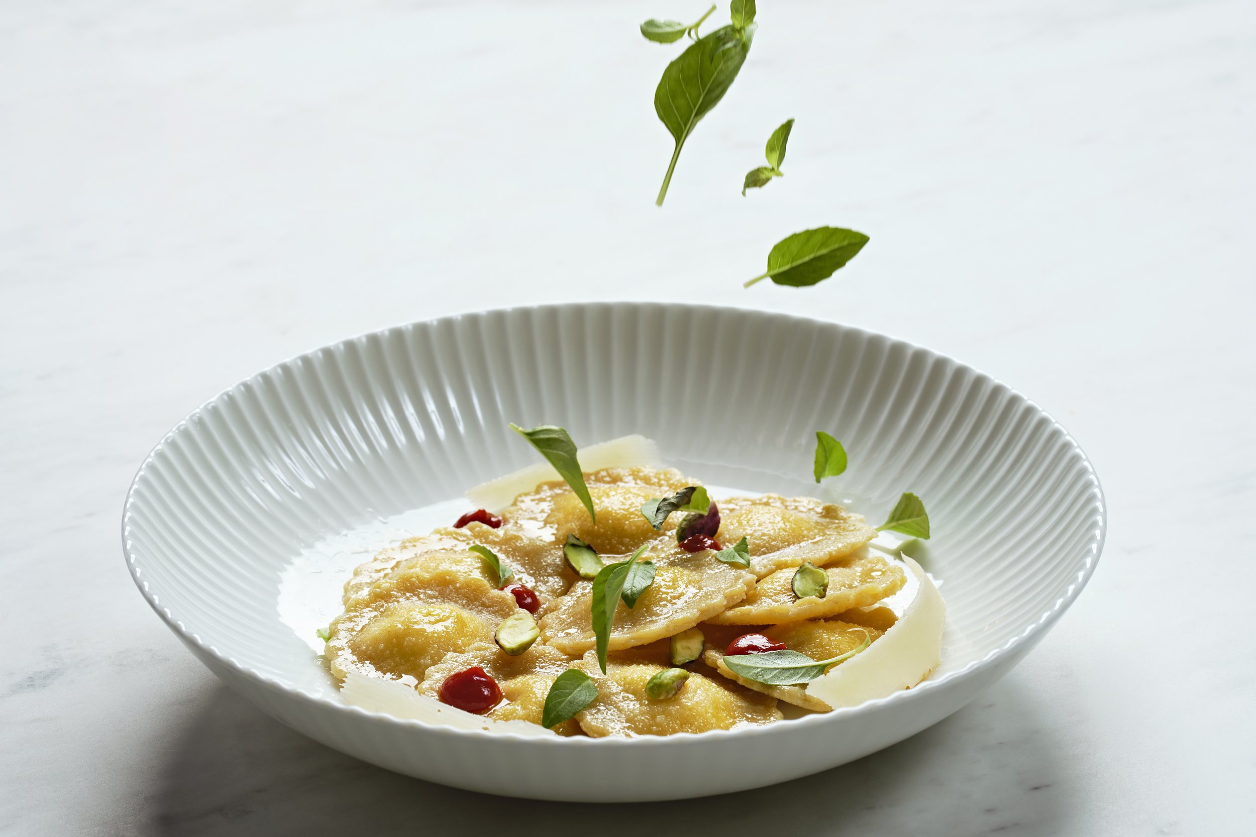 A white textured bowl contains ravioli pasta garnished with green leaves, sliced pistachios, and red condiment or sauce, with some leaves falling onto the bowl.