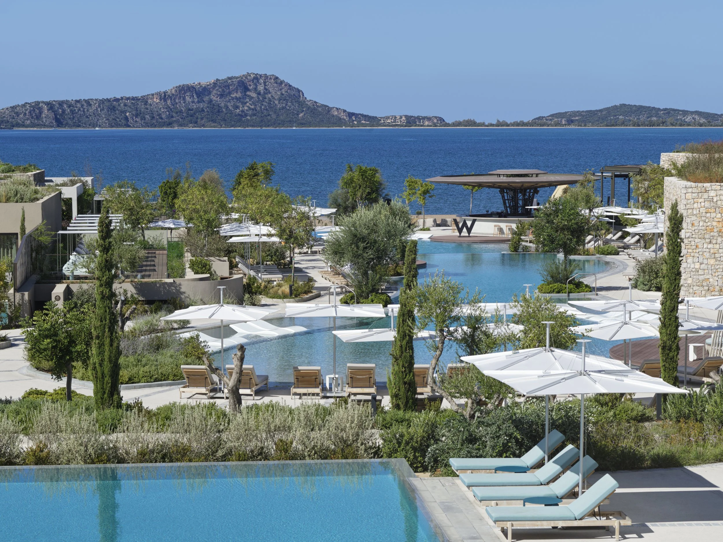 Luxury resort pool area with lounge chairs, umbrellas, and trees overlooking a lake or seaside with hills in the background.