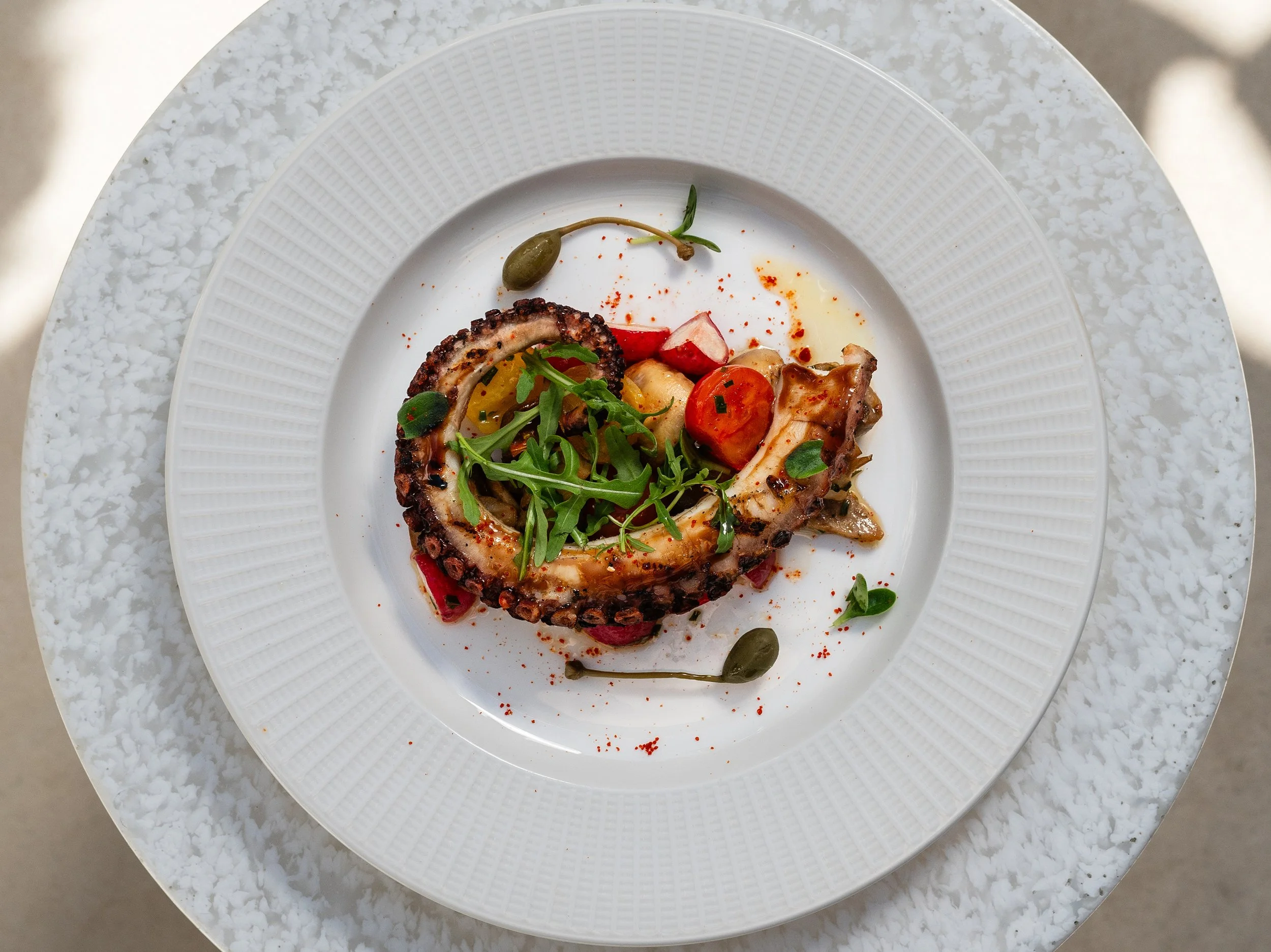 A white plate with grilled octopus top, garnished with arugula, cherry tomatoes, capers, and radishes, on a white textured surface.