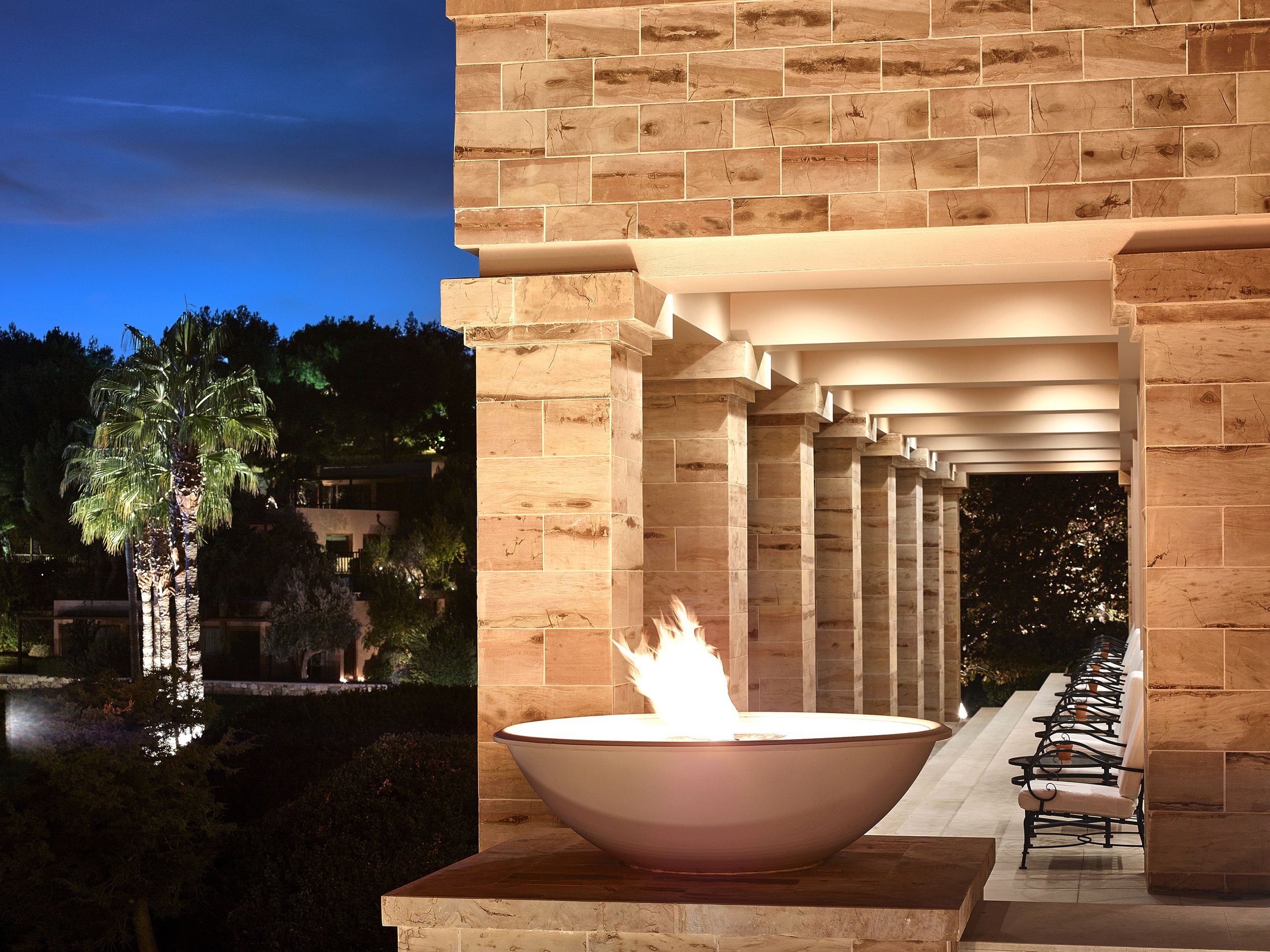 Nighttime view of a modern outdoor patio with a fire bowl and seating area, featuring stone columns and roof, surrounded by trees and illuminated palm trees in the background.