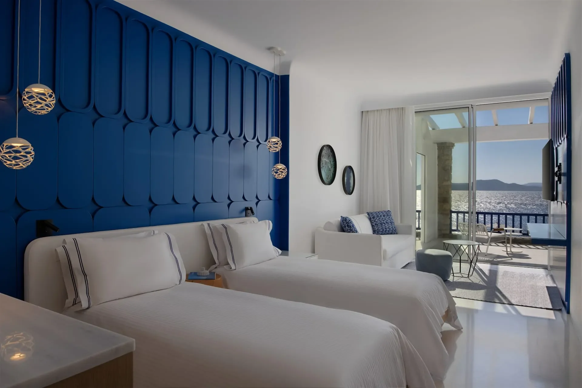 A modern hotel room with two beds, white bedding, blue accent wall with decorative panels, hanging pendant lights, a white sofa with blue pillows, a small table, and a sliding glass door opening to a balcony overlooking a body of water and distant mo