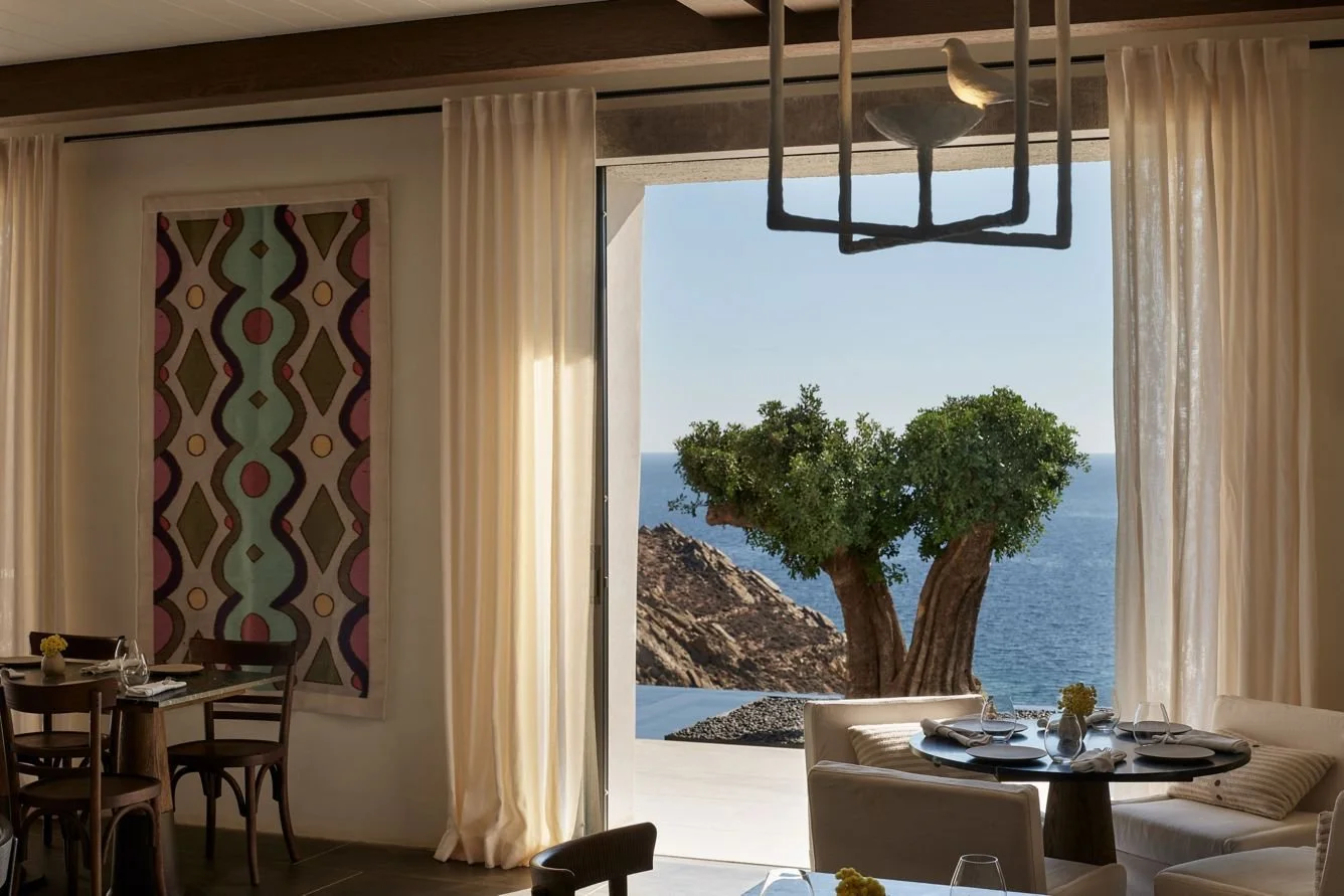 Interior of a seaside restaurant with a large window viewing a tree and ocean, featuring dining tables, chairs, and modern decor.