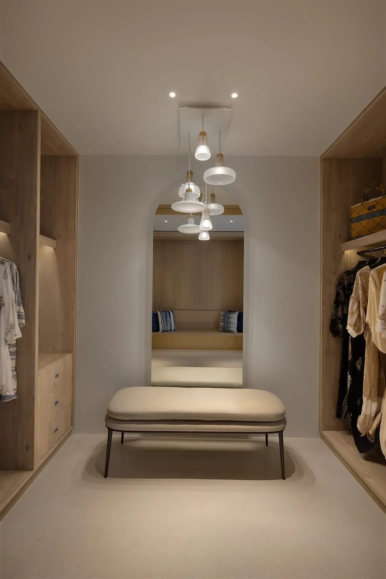 A walk-in closet with built-in wooden shelving on both sides, a cushioned bench in the middle, and a mirror at the end. There's a modern pendant light fixture hanging from the ceiling.