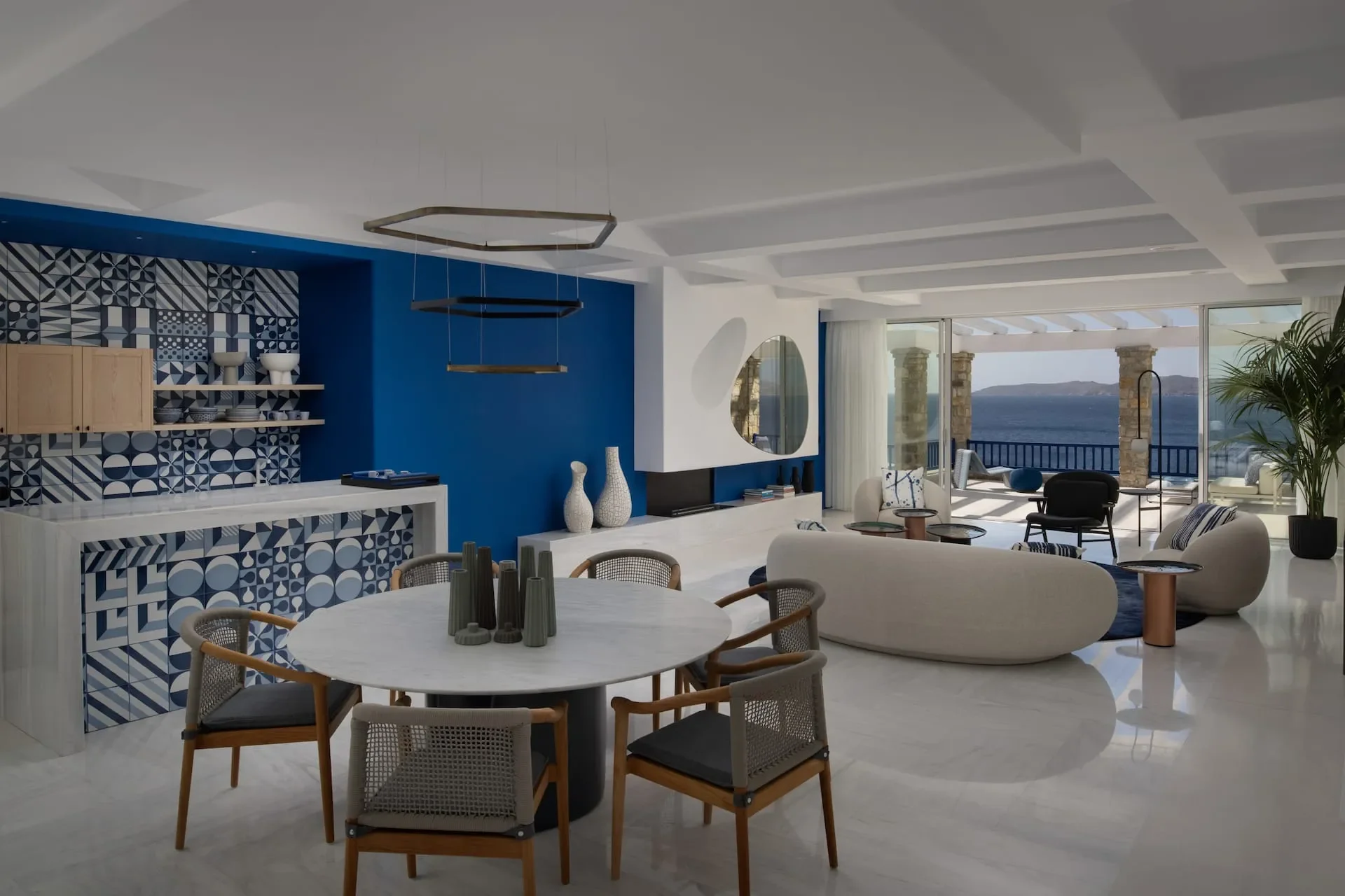 Open-concept living and dining area overlooking an ocean, with white and blue decor, modern furniture, a round marble dining table, and large sliding glass doors leading to an outdoor patio.