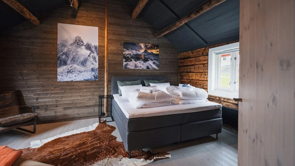 A cozy bedroom with wooden walls and ceiling, featuring a bed with pillows and blankets, a window with daylight, mountain landscape paintings on the wall, a small armchair, and a brown and white fur rug on the light-colored wooden floor.