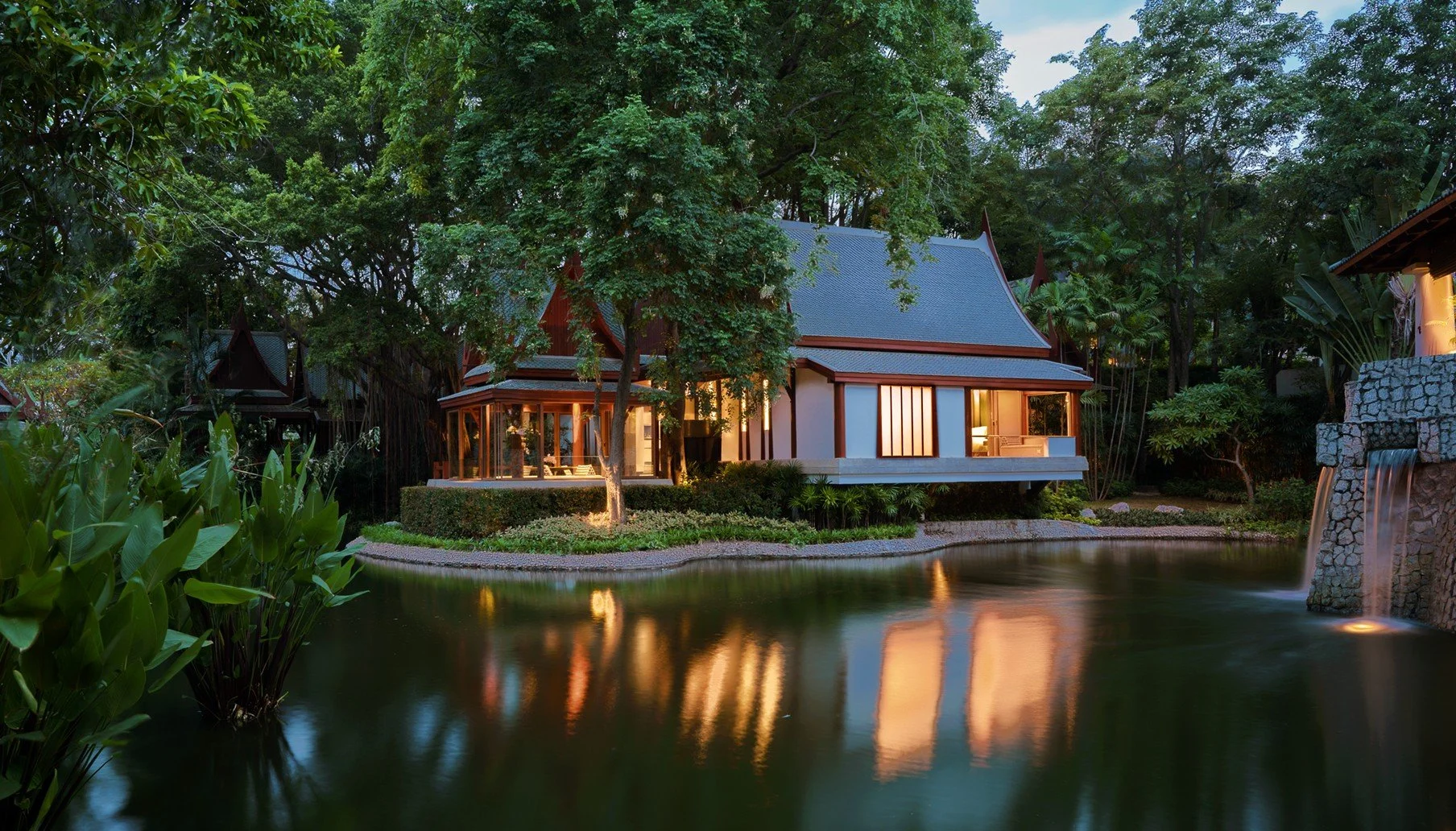 A traditional style house with a blue roof and white walls, surrounded by dense green trees, situated near a calm pond with reflections of the house and trees, during dusk with interior lights on.