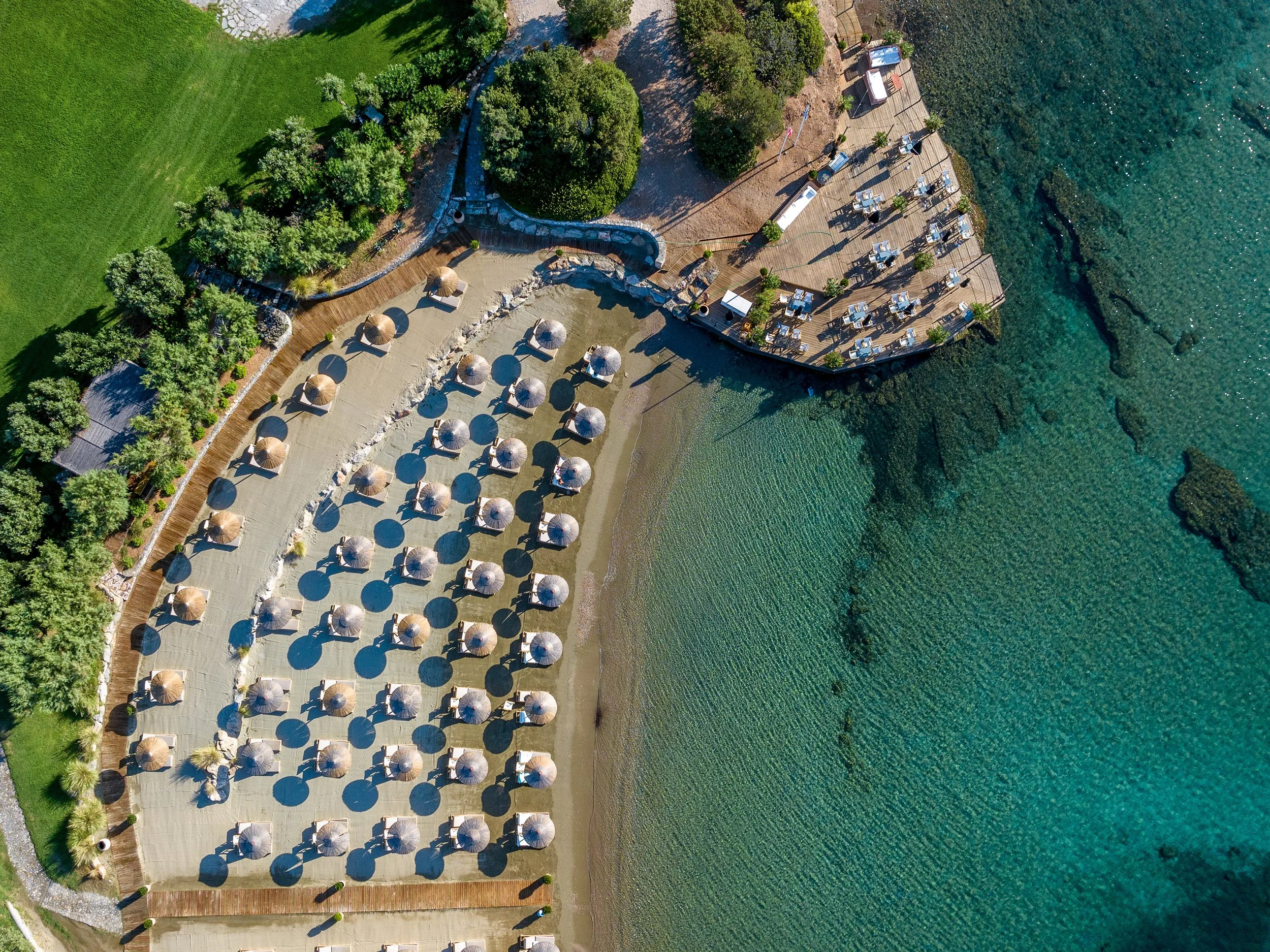 Aerial view of a beach with umbrellas, a wooden deck with tables and chairs, surrounded by green trees and grass, near a body of clear blue water.