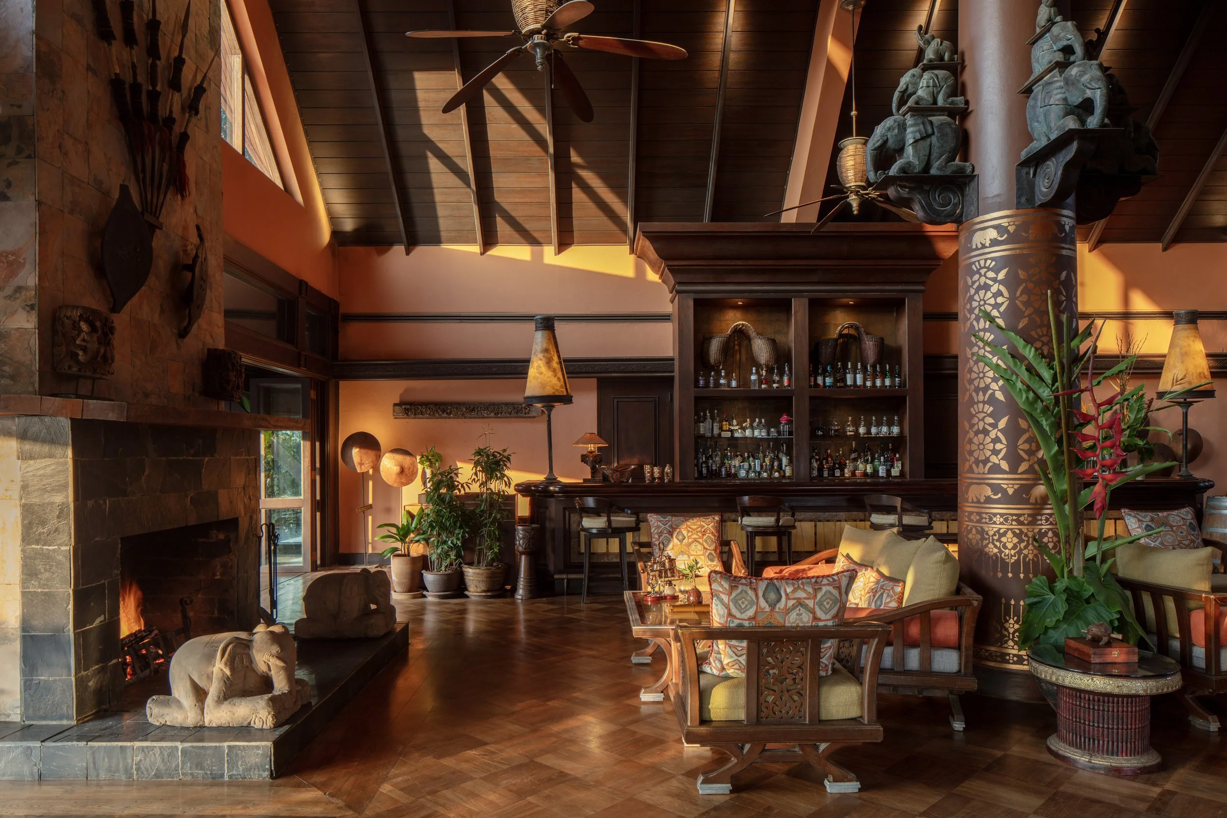 A cozy living room with a high wooden ceiling, a stone fireplace with a carved elephant statue, potted plants, colorful cushions, a bar with bottles on shelves, and tropical decor accents.