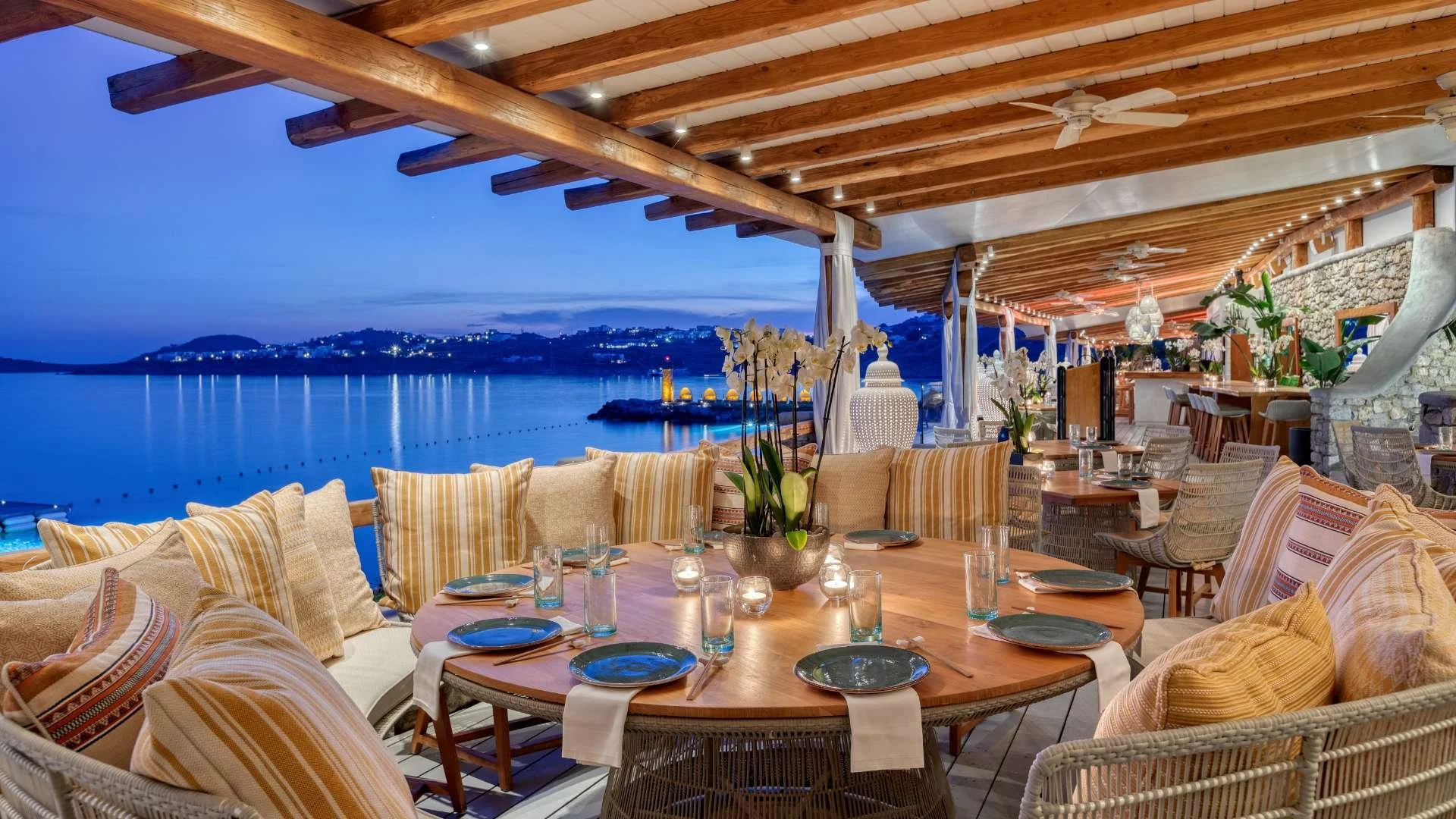 A beachfront restaurant with a large round dining table set with plates, glasses, candles, and a floral centerpiece, overlooking a calm body of water and a distant hillside town illuminated at dusk.