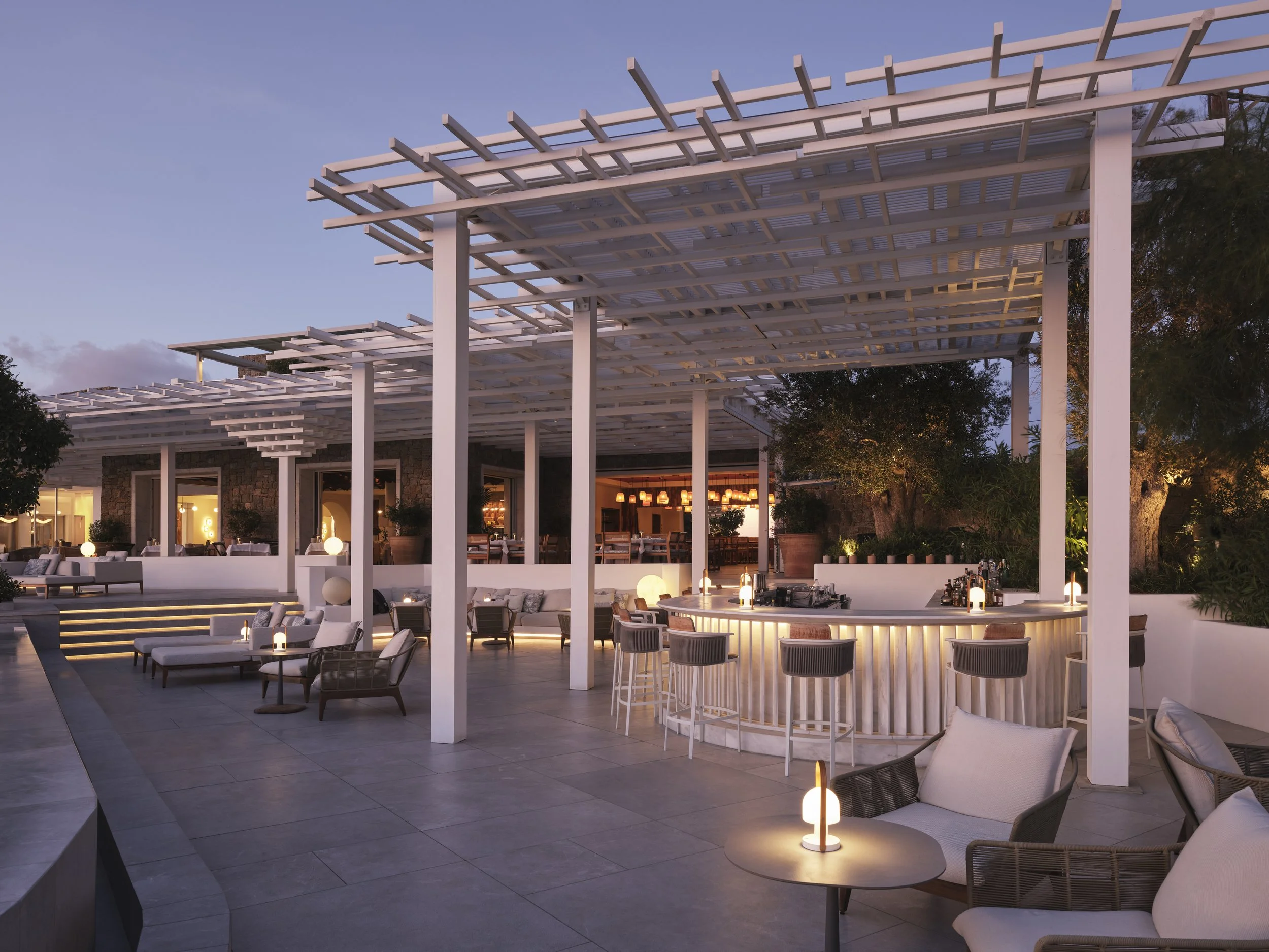 Outdoor patio area with seating and bar under a white pergola at sunset, featuring lounge chairs, bar stools, and ambient lighting.