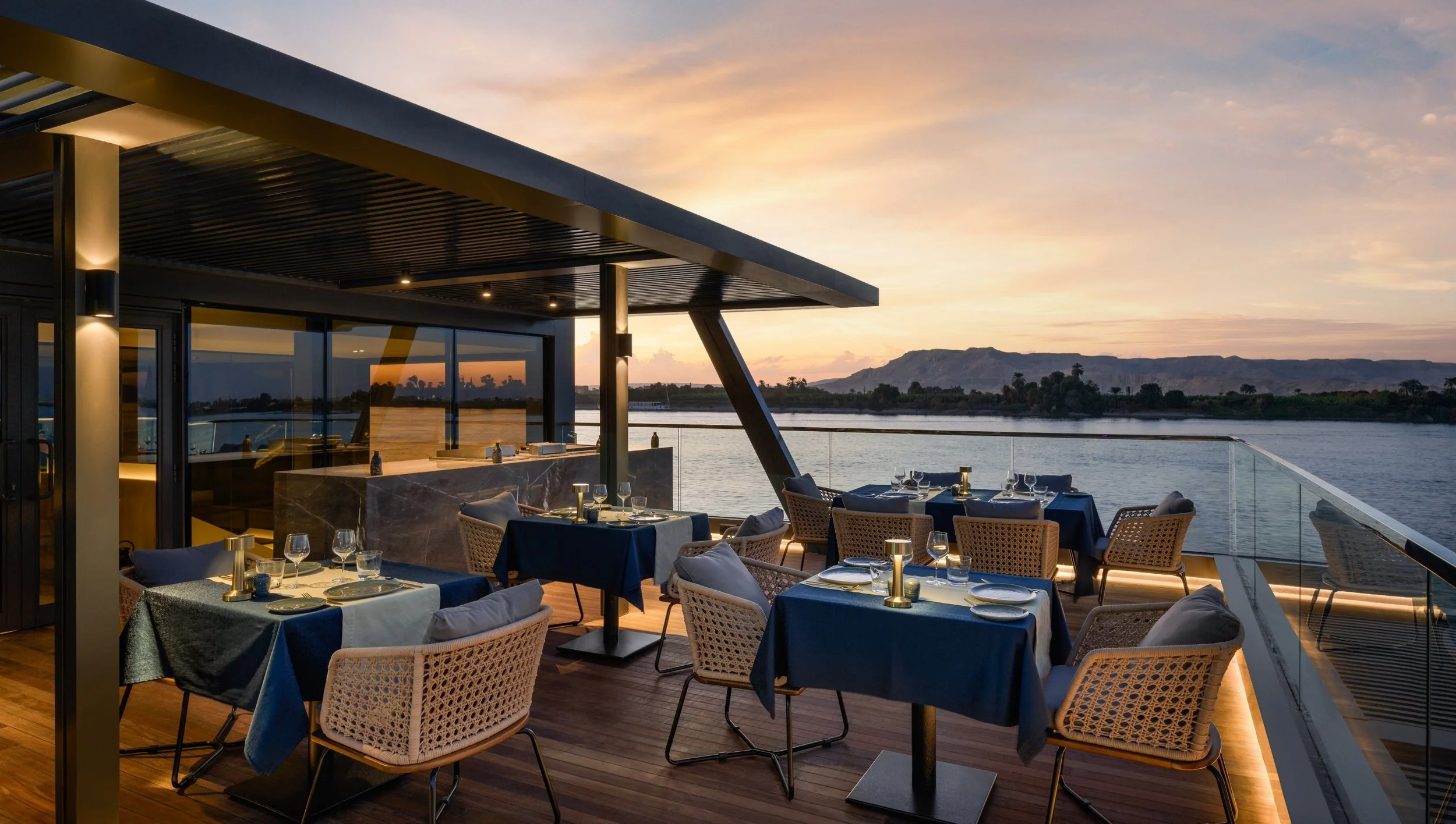 Outdoor restaurant on a boat or waterfront during sunset, with tables set with blue tablecloths, wine glasses, and silverware, overlooking water and distant hills.