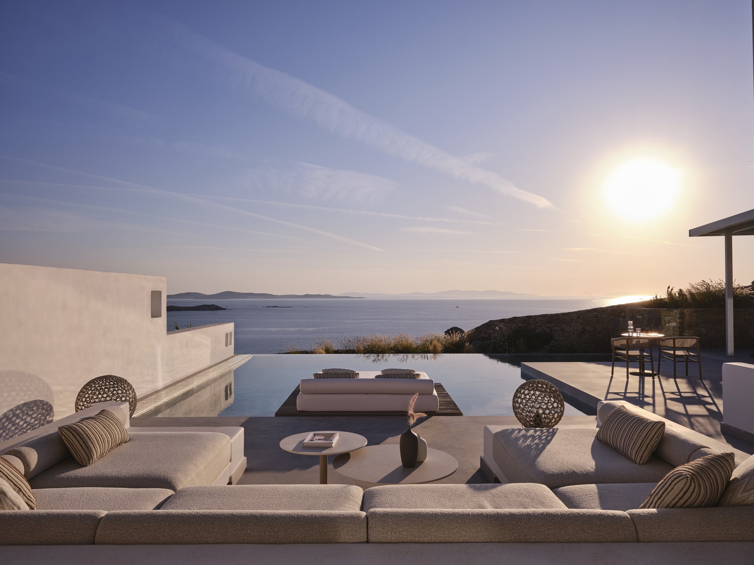 A luxurious outdoor patio with a view of the ocean, featuring a sitting area with beige sofa and cushions, a table with a book and decoration, an infinity pool, and a small table with chairs, all illuminated by the setting sun.