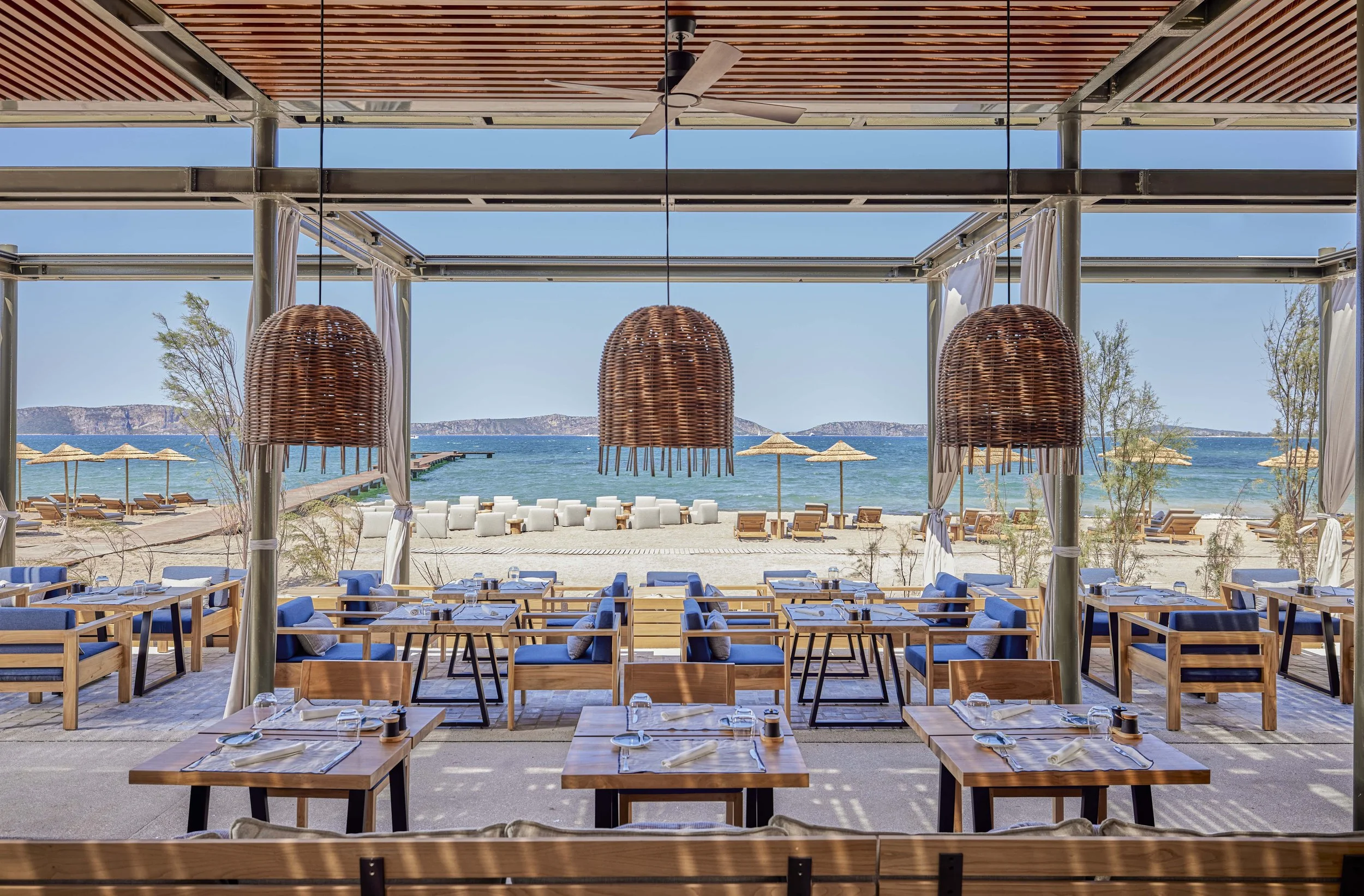 An open-air beachfront restaurant with wooden tables and blue cushioned chairs, overlooking a sandy beach with lounge chairs and umbrellas, and the ocean beyond under a clear blue sky.