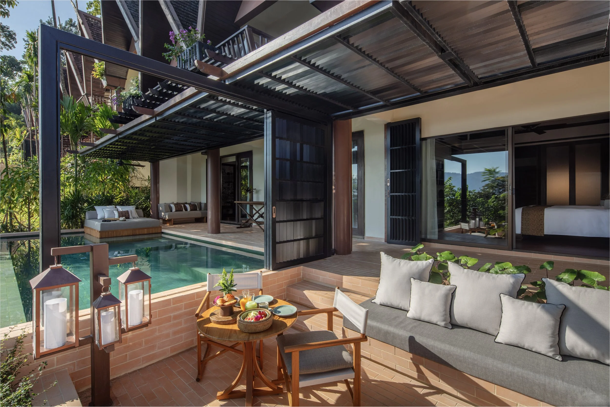 Outdoor patio area with a small pool, seating cushions on a lounge, a table set with breakfast, and a shaded sectional sofa, overlooking lush greenery and mountains in the background.