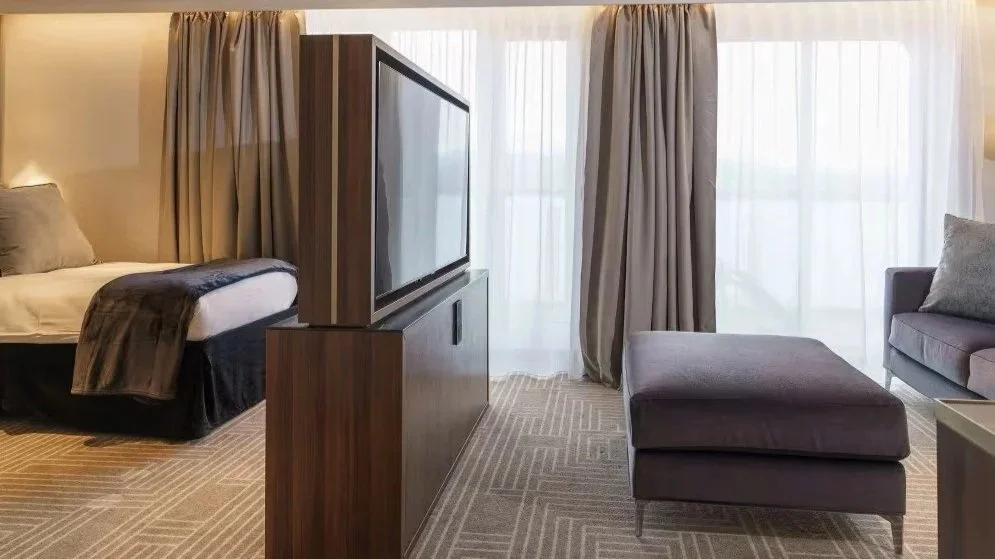 A modern hotel room with a bed on the left, a TV on a wooden stand in the middle, and a grey chaise lounge with a matching sofa and pillows near large windows with beige curtains.