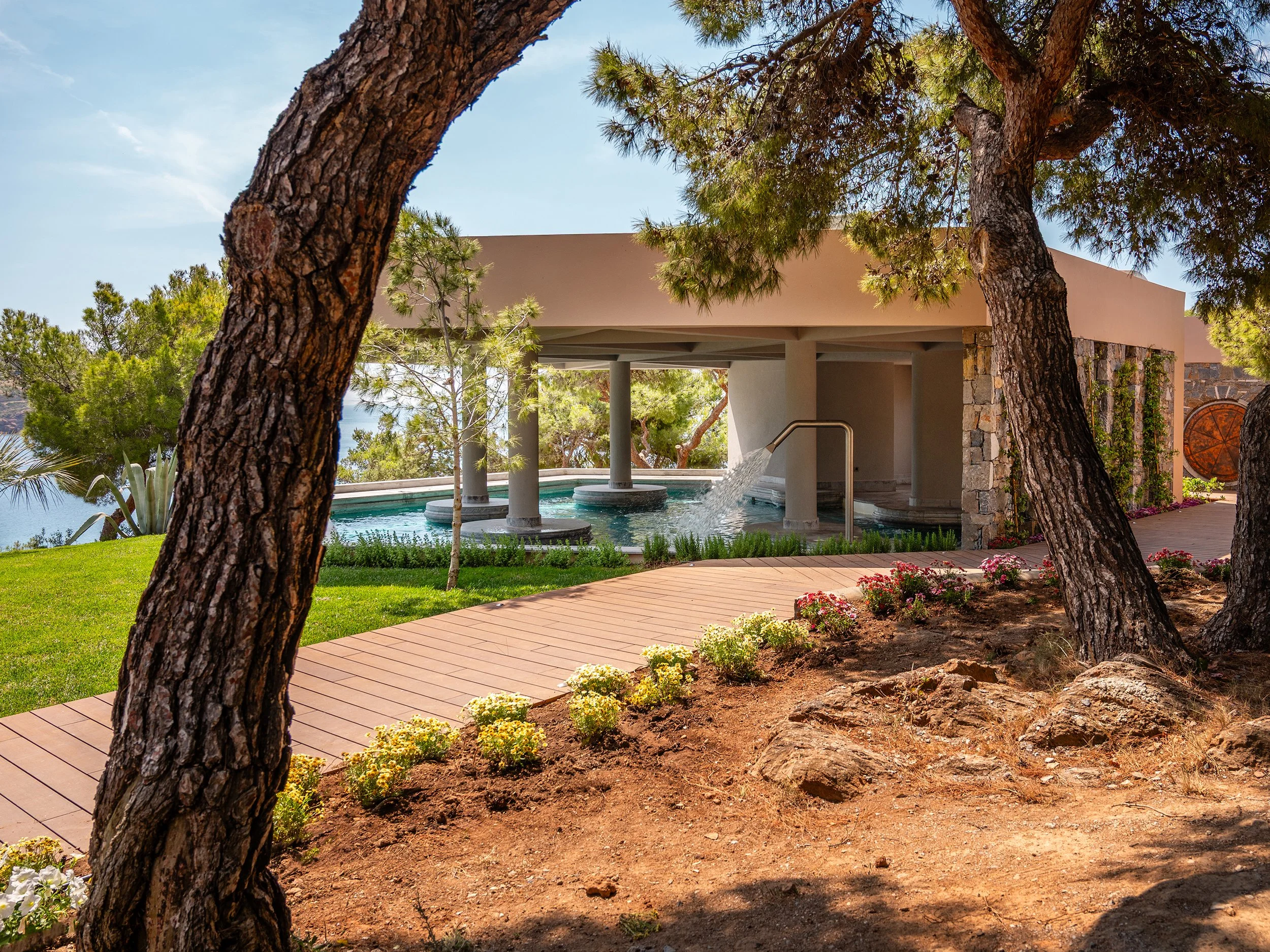 A modern outdoor pool with waterfalls and a covered area, surrounded by trees and flowering plants, with a view of water in the background.