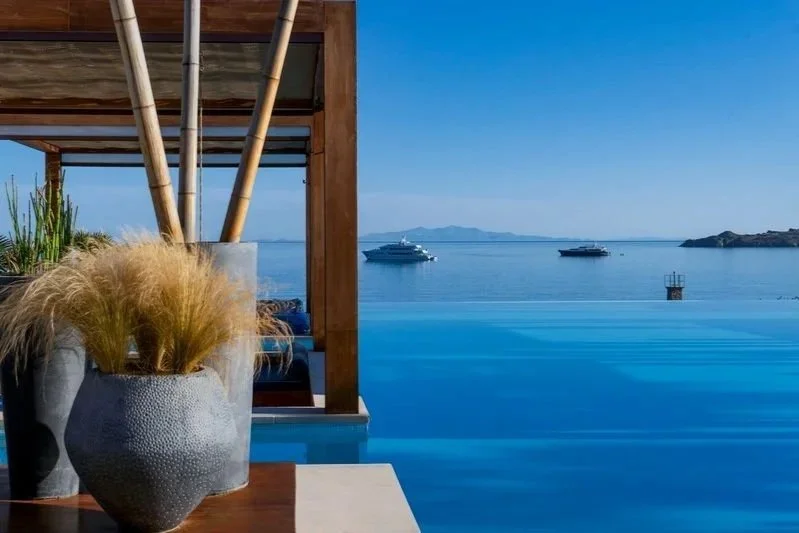 View of an infinity pool overlooking calm ocean water with boats anchored in the distance, and rocky islands under a clear blue sky, with part of a wooden structure and potted plants in the foreground.