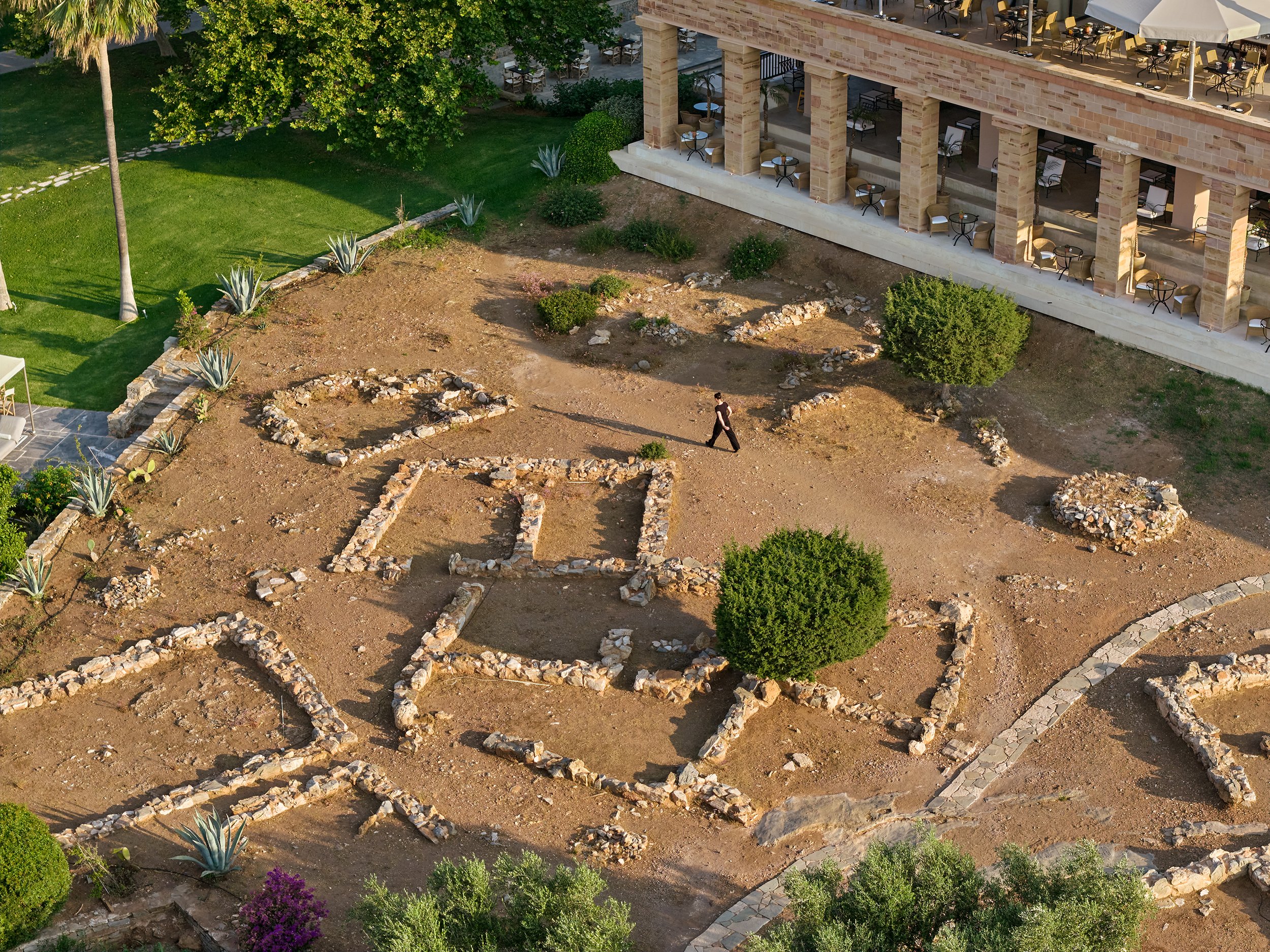 Aerial view of a landscaped area with stone outlines on the ground forming various shapes, including a cross and other geometric figures. There are green bushes, a few trees, and some plants, with a building featuring a balcony or terrace with tables