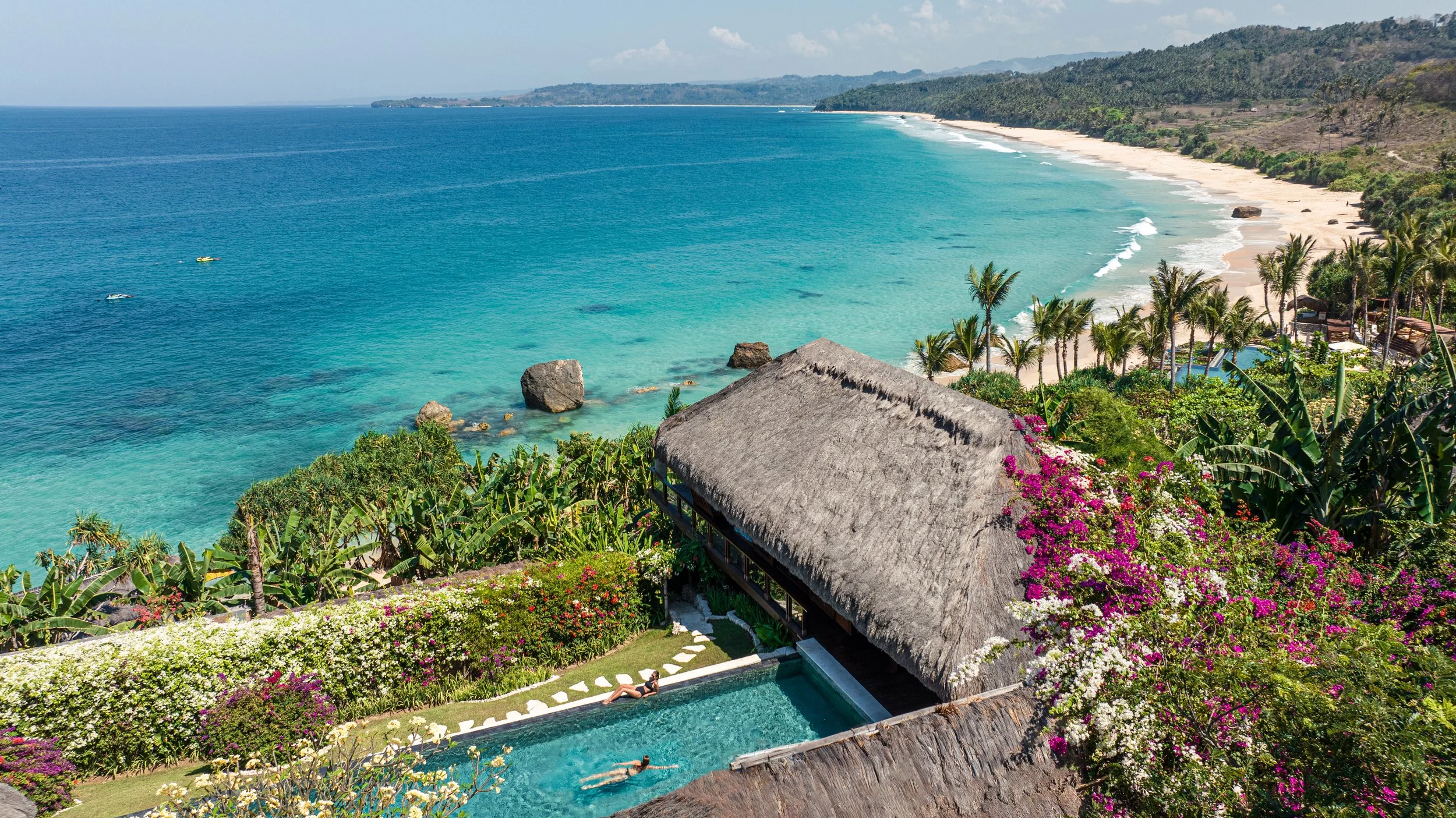 A tropical beach with turquoise water, palm trees, and lush greenery. In the foreground, there is a thatched-roof hut and a swimming pool with people swimming, surrounded by vibrant flowers.