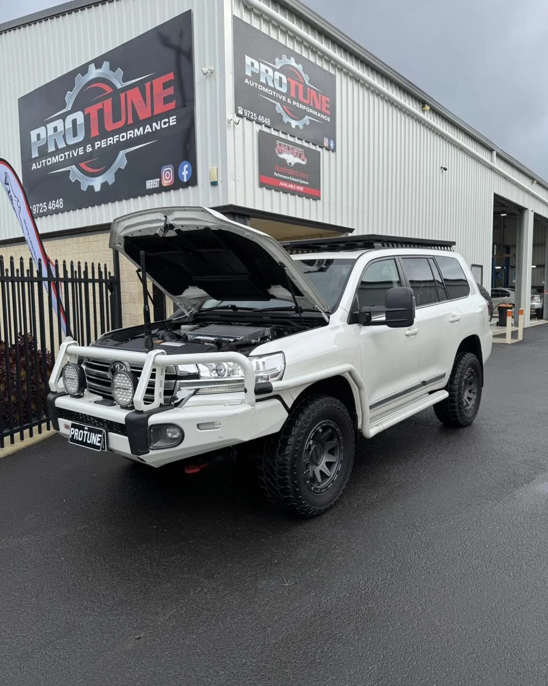 White SUV with its hood open, parked in front of an automotive performance shop called Pro Tune, which has banners and signs on the building.