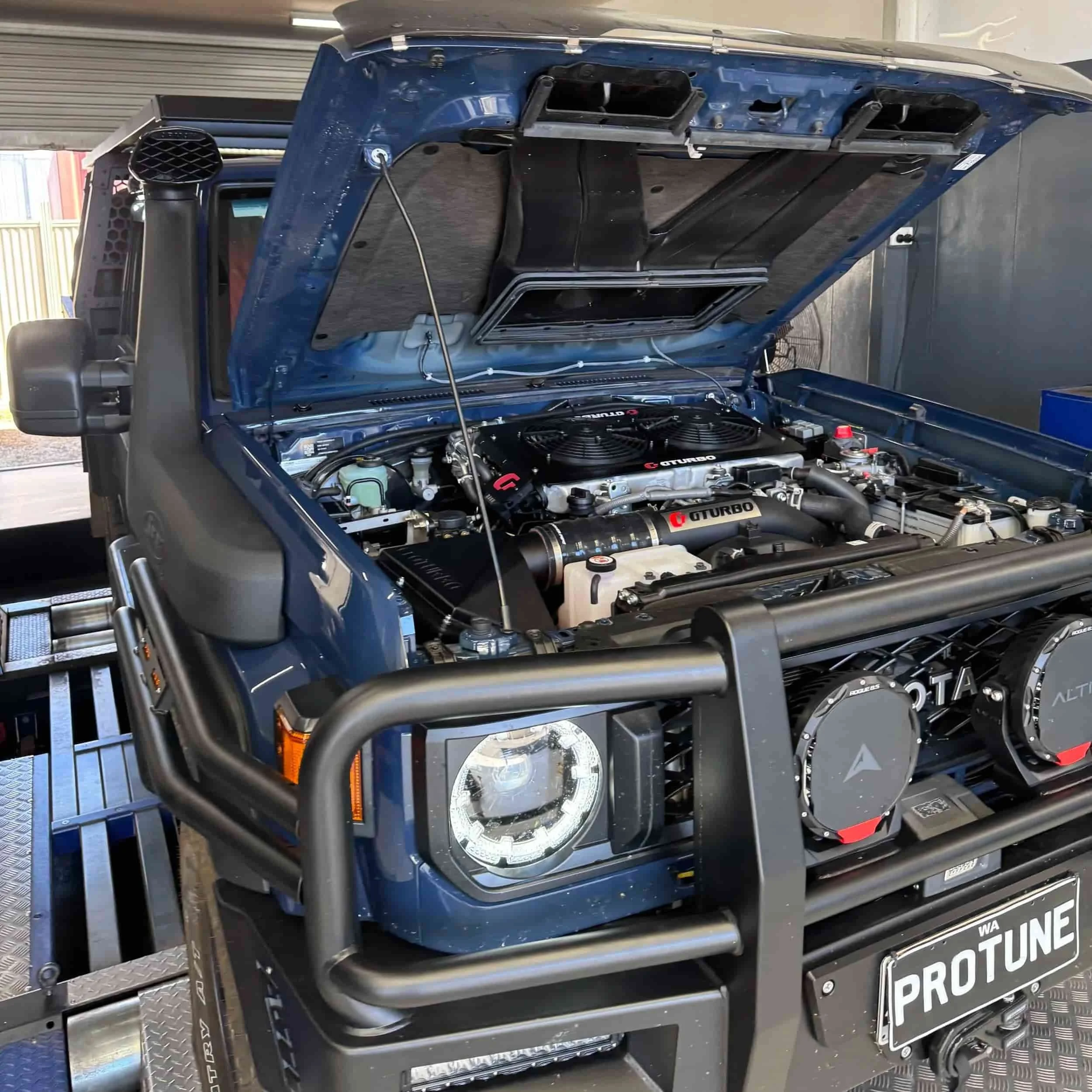 Open hood of a blue off-road vehicle showing a high-performance turbocharged engine and front grille with headlights, parked inside a garage.
