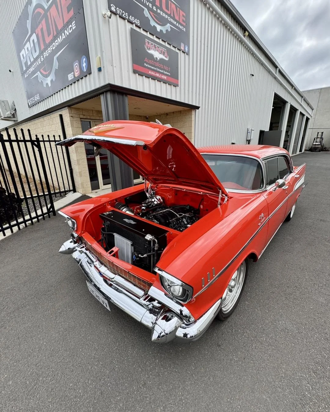 A vintage Chevrolet car painted red with a chrome bumper, parked outside a building with a sign for automotive performance shop. The hood is open, revealing a clean engine bay and engine components.